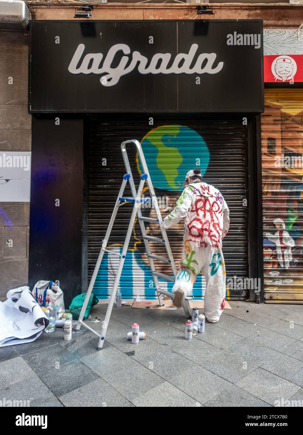 Un artista di strada con una tuta dipinta che crea un murale colorato su una tapparella di Siviglia, in Spagna. Foto Stock