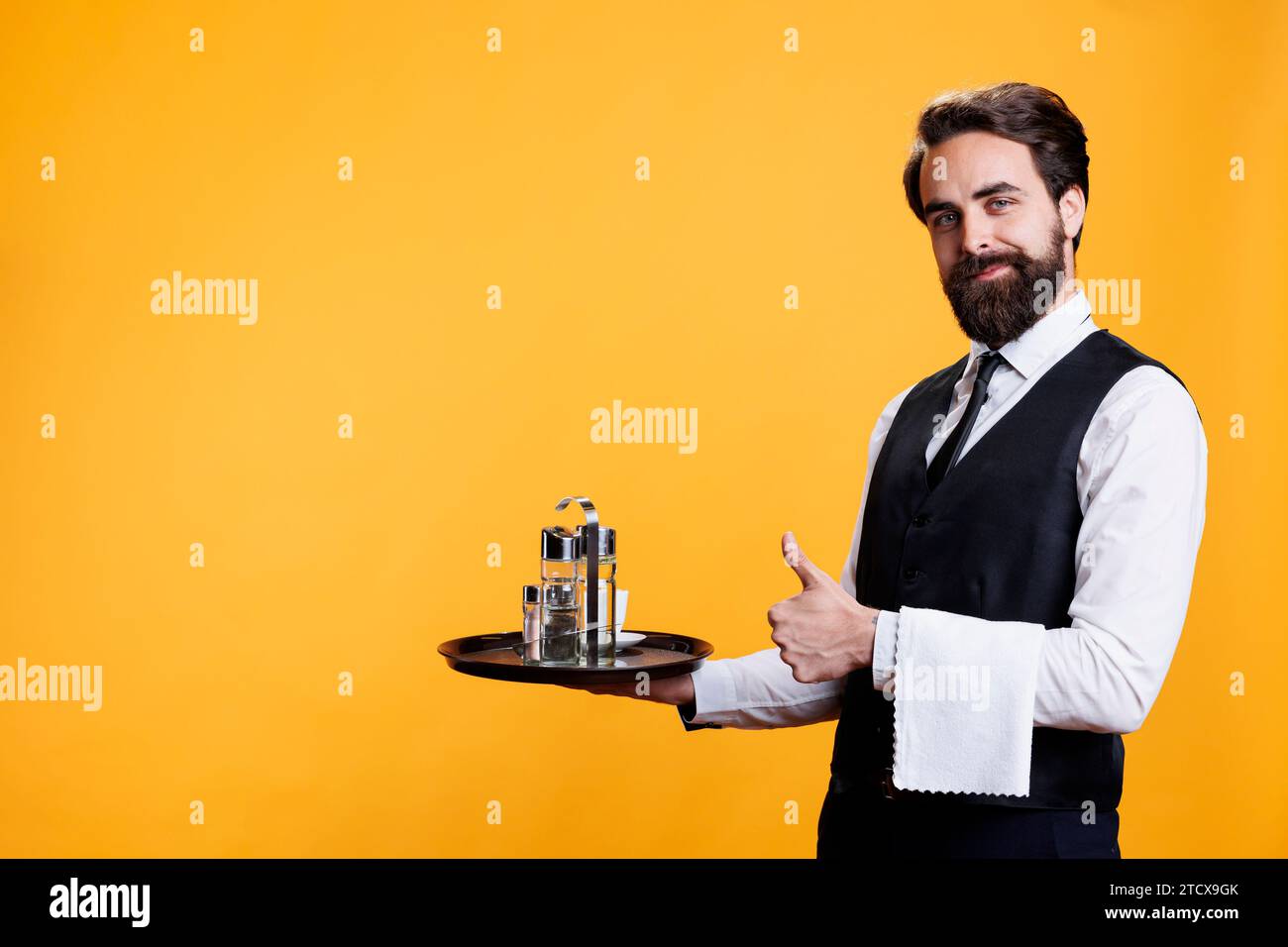 Il maggiordomo sorridente ama registrarsi in studio per esprimere positività e approvazione con il simbolo del pollice in alto. Elegante e felice operaio del ristorante presenta un gesto d'accordo sullo sfondo giallo. Foto Stock