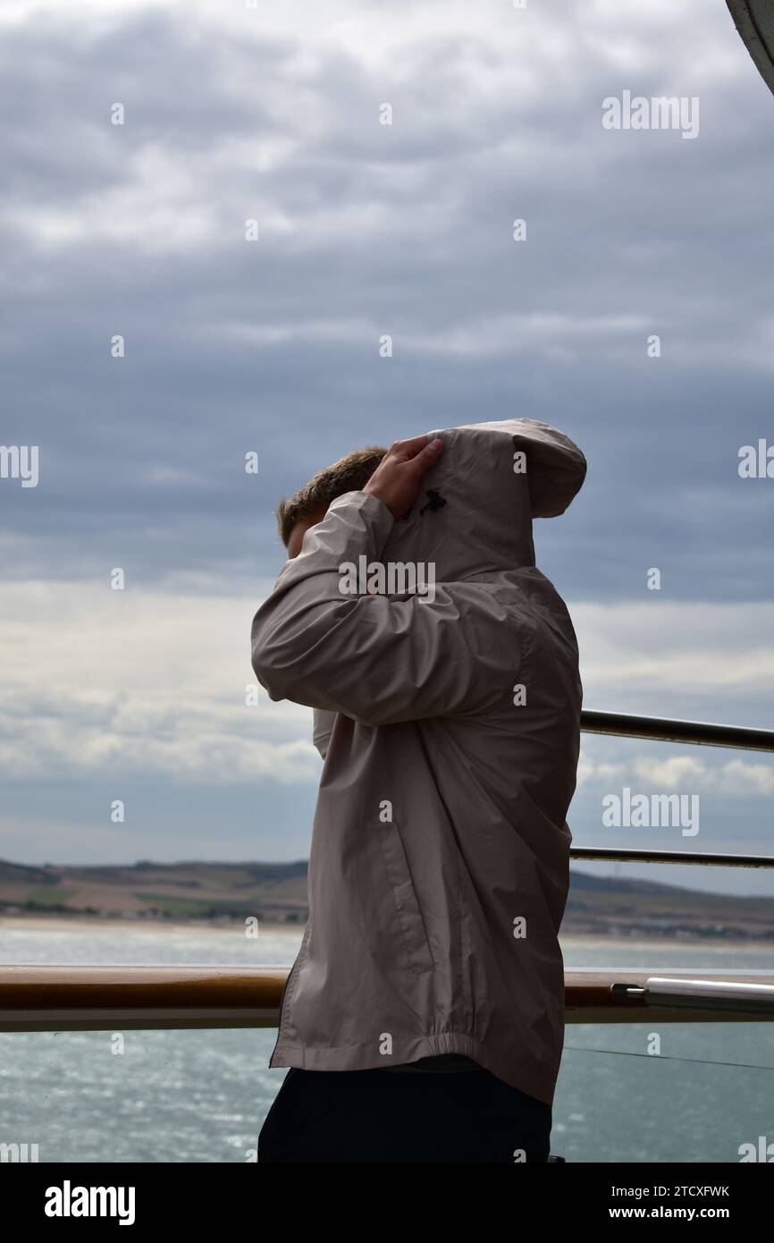 Giovane uomo con pantaloni neri e giacca grigia che rimuove il cappuccio della giacca impermeabile mentre si trova accanto a un corrimano su una nave a vela vicino alla costa Foto Stock