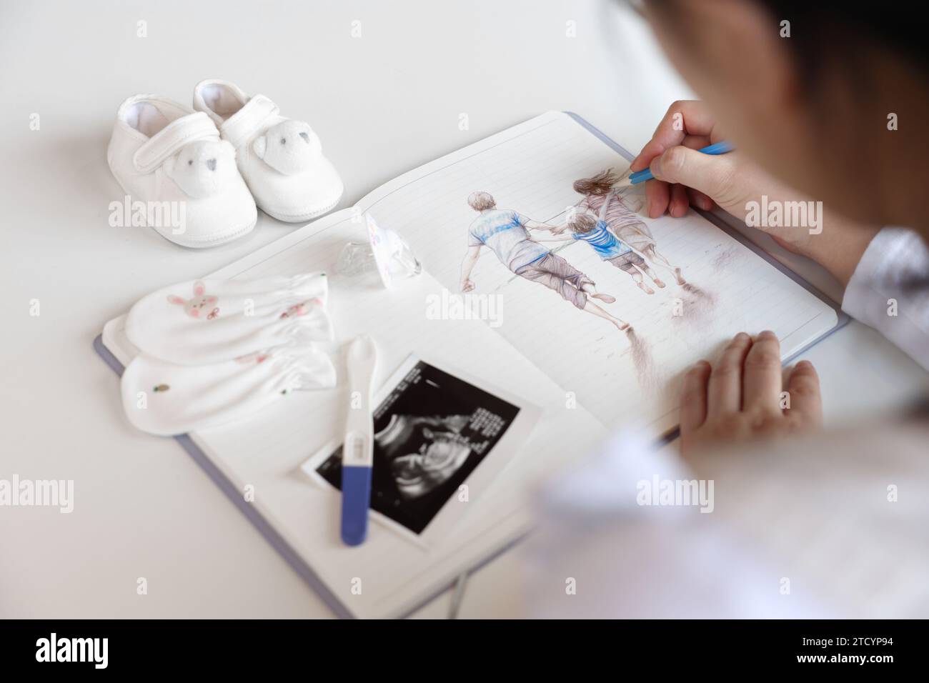 Una donna incinta disegna l'immagine della sua famiglia in un notebook con una matita, vengono posizionati oggetti per bambini e immagini ecografiche Foto Stock