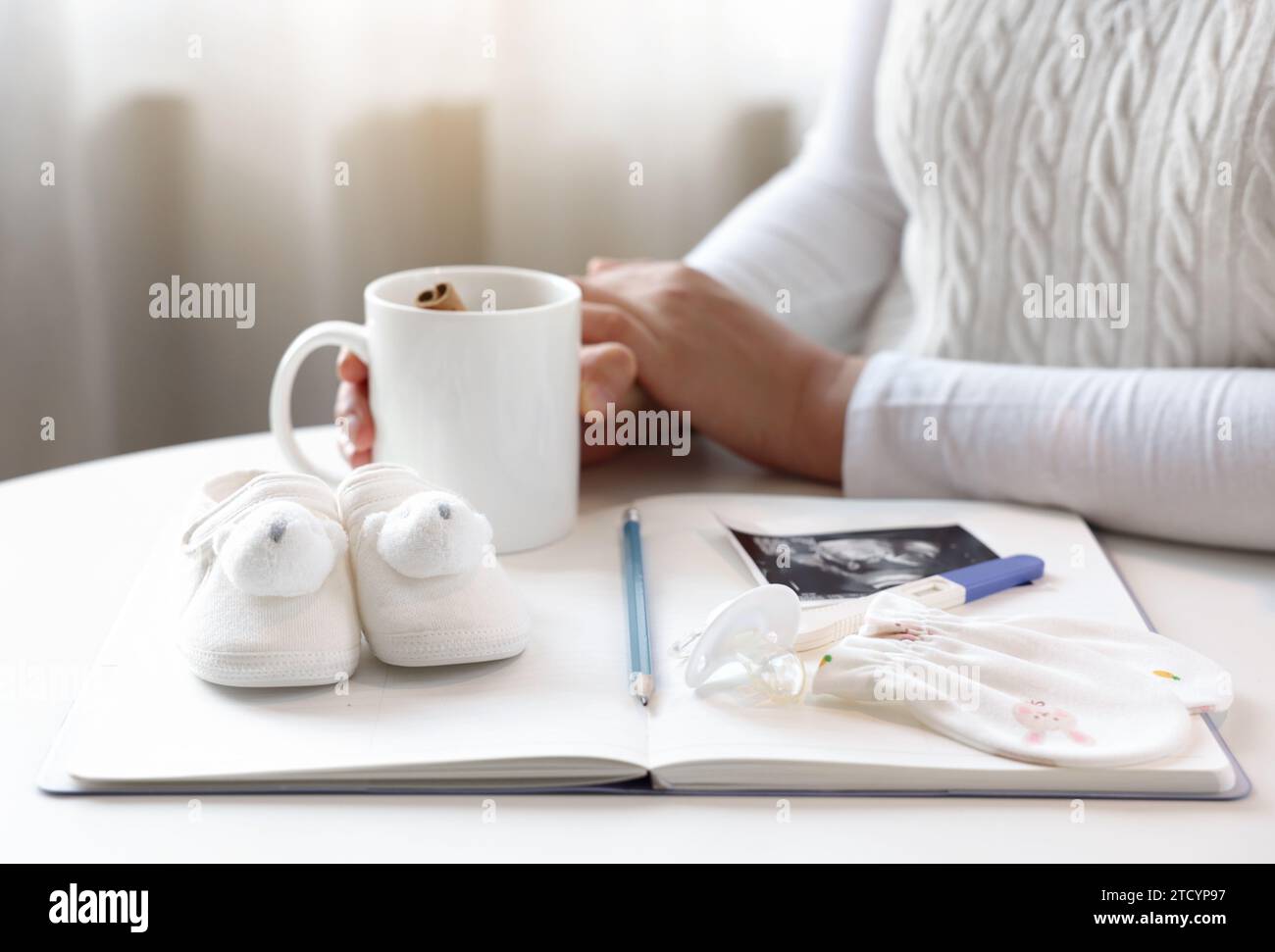 Una donna incinta ti aspetta con una tazzina da tè, davanti a te notebook e oggetti per bambini Foto Stock