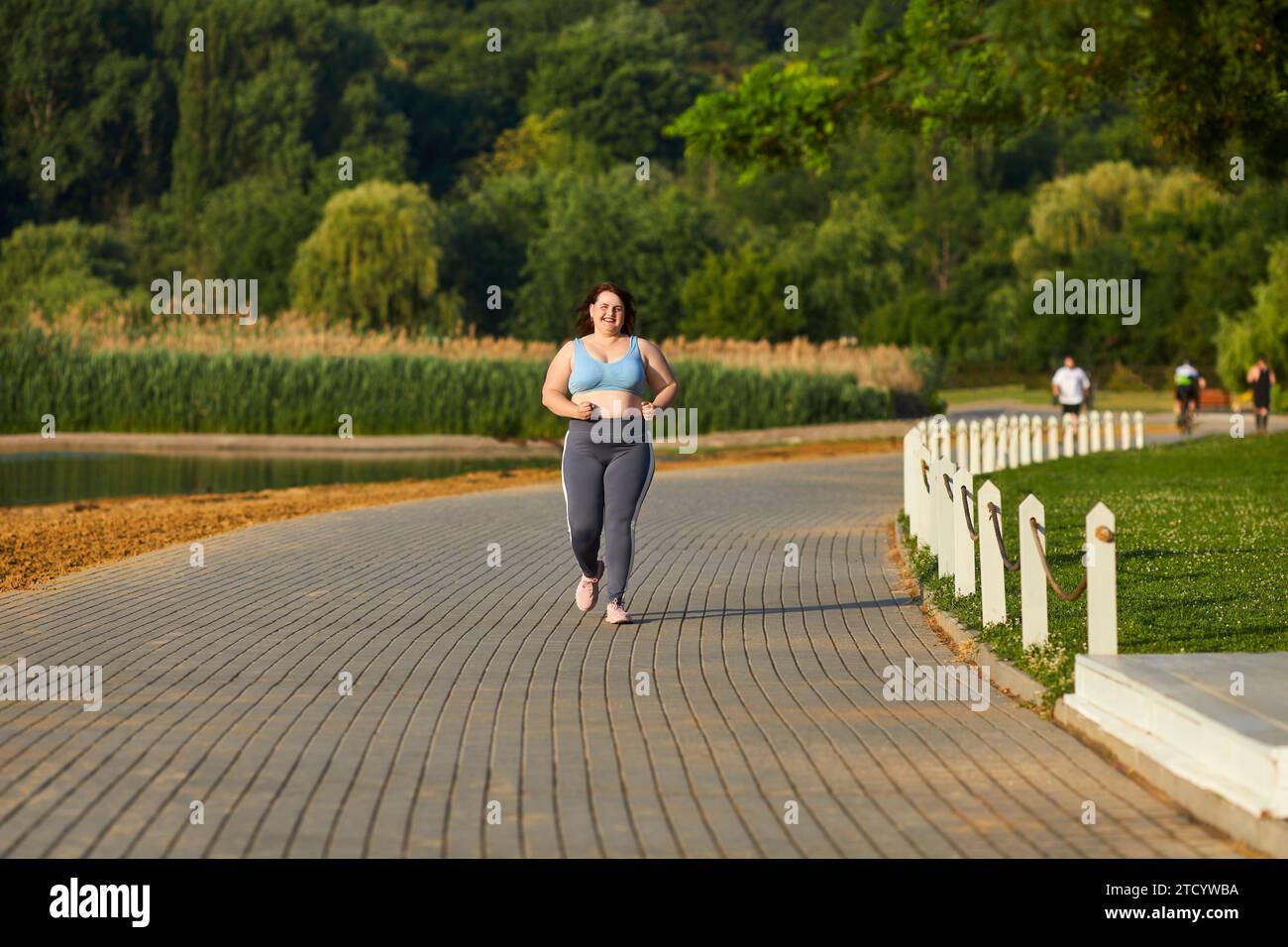 Donna grassa e sorridente in sovrappeso che corre nel parco estivo in mezzo alla natura. Foto Stock