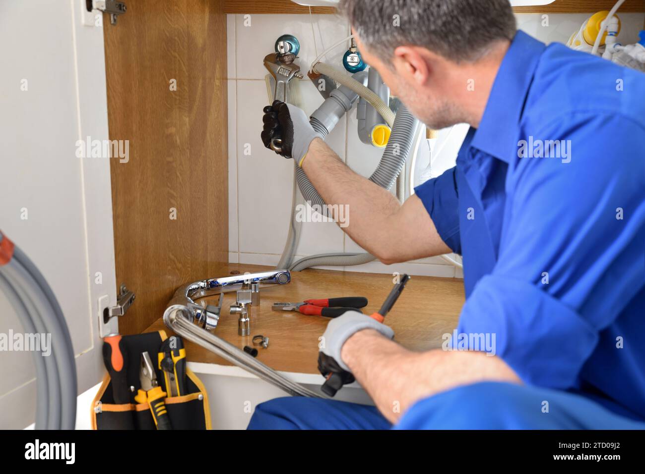 Particolare dell'idraulico in uniforme riparando una perdita sotto il lavandino in una casa con gli attrezzi intorno. Composizione orizzontale. Foto Stock