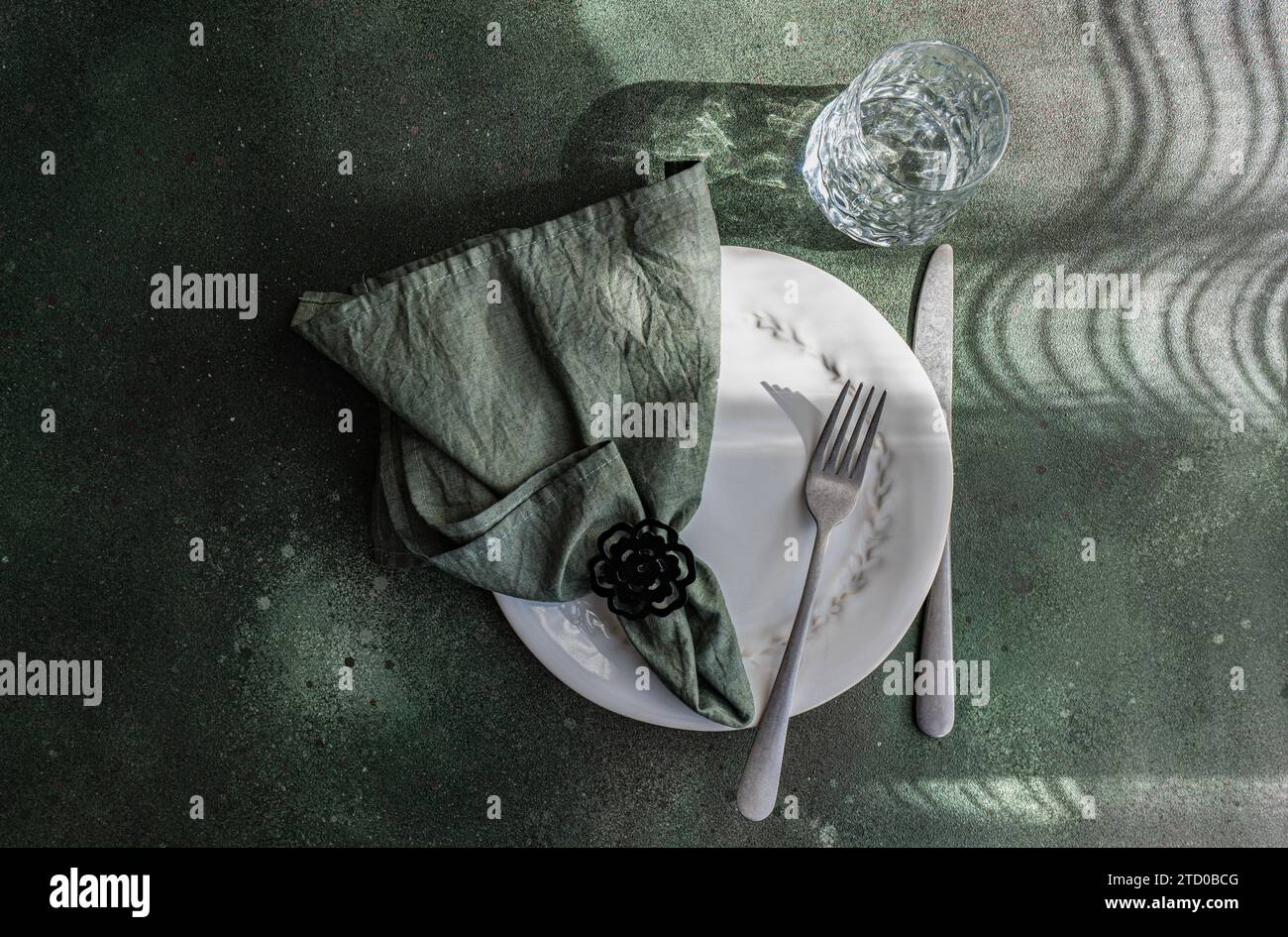 Un sofisticato tavolo con un piatto bianco, posate, un tovagliolo con un anello e un vetro che fonde ombre e riflessi giocosi. Foto Stock