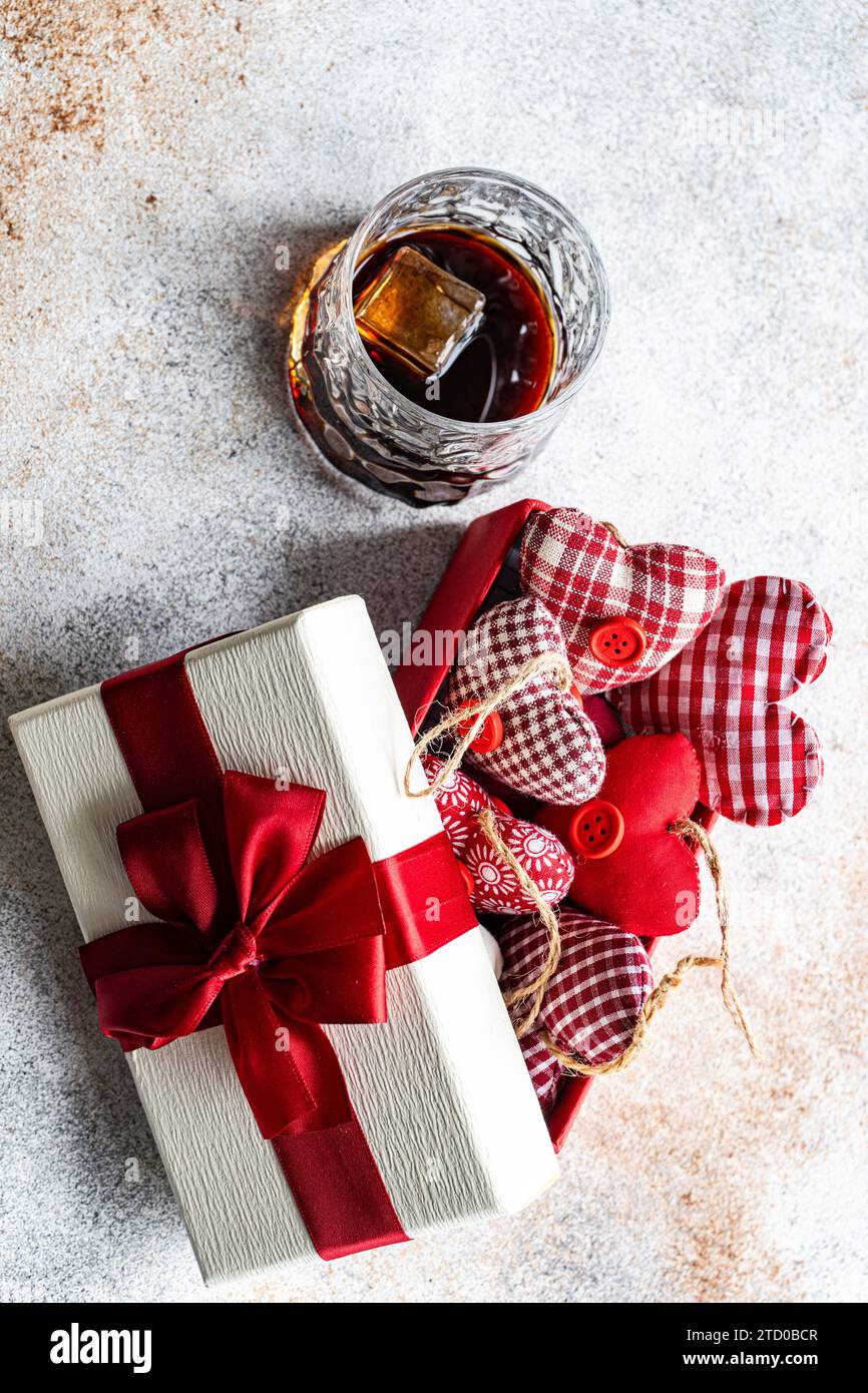 Elegante confezione regalo con un nastro rosso, ornamenti a forma di cuore e un bicchiere di whisky, che evocano un'atmosfera celebrativa. Foto Stock