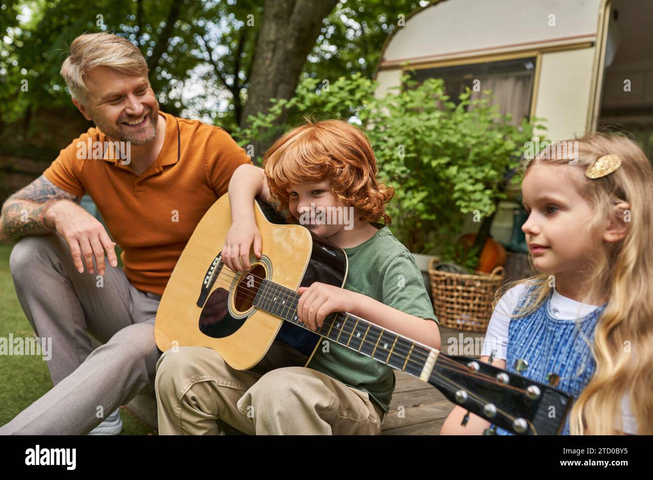 redhead boy che suona la chitarra vicino a padre e sorella sorridenti accanto a casa trailer, attività creative Foto Stock