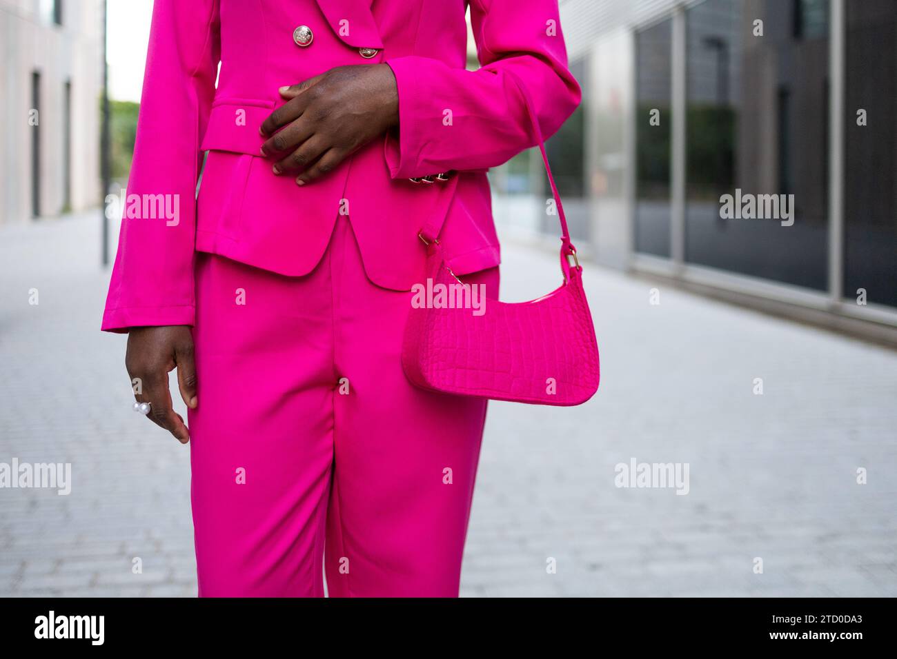 Ritaglia una donna afroamericana anonima con un vestito rosa e una borsa che cammina su una strada lastricata con pareti di vetro riflettenti contro lo sfondo sfocato Foto Stock