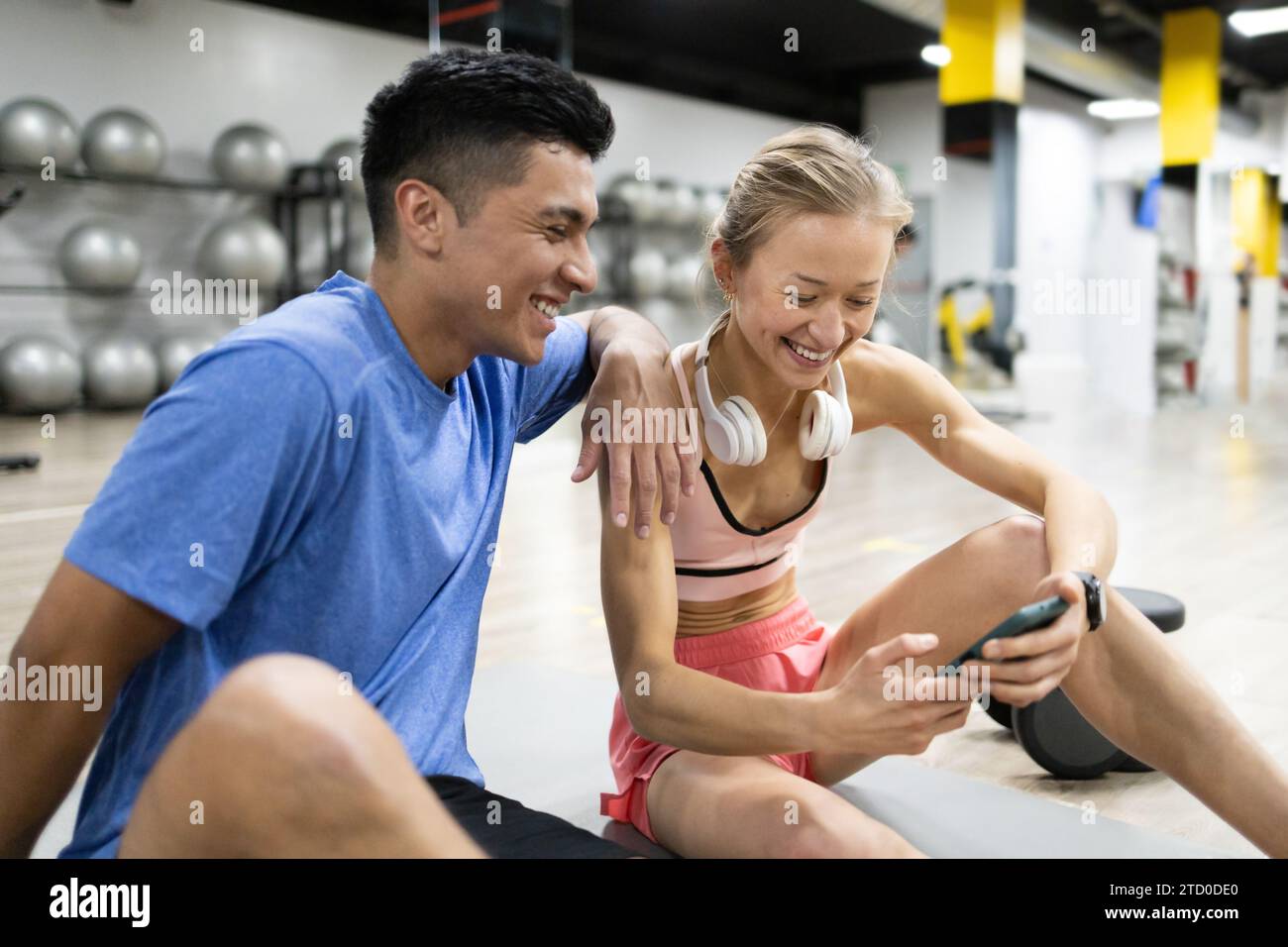 Due appassionati di fitness fanno una pausa in palestra, ridendo insieme mentre guardano uno smartphone con le palle da ginnastica sullo sfondo. Foto Stock