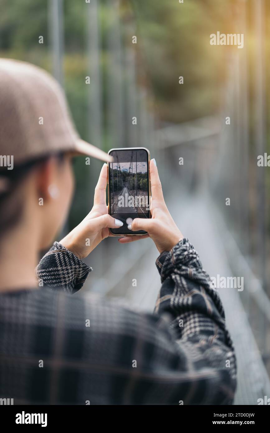 Ritagliate la vista di un'escursionista anonima che indossa un berretto e delle casuali che fotografano la passerella pedonale con lo smartphone durante il trekking nella foresta durante il fine settimana Foto Stock