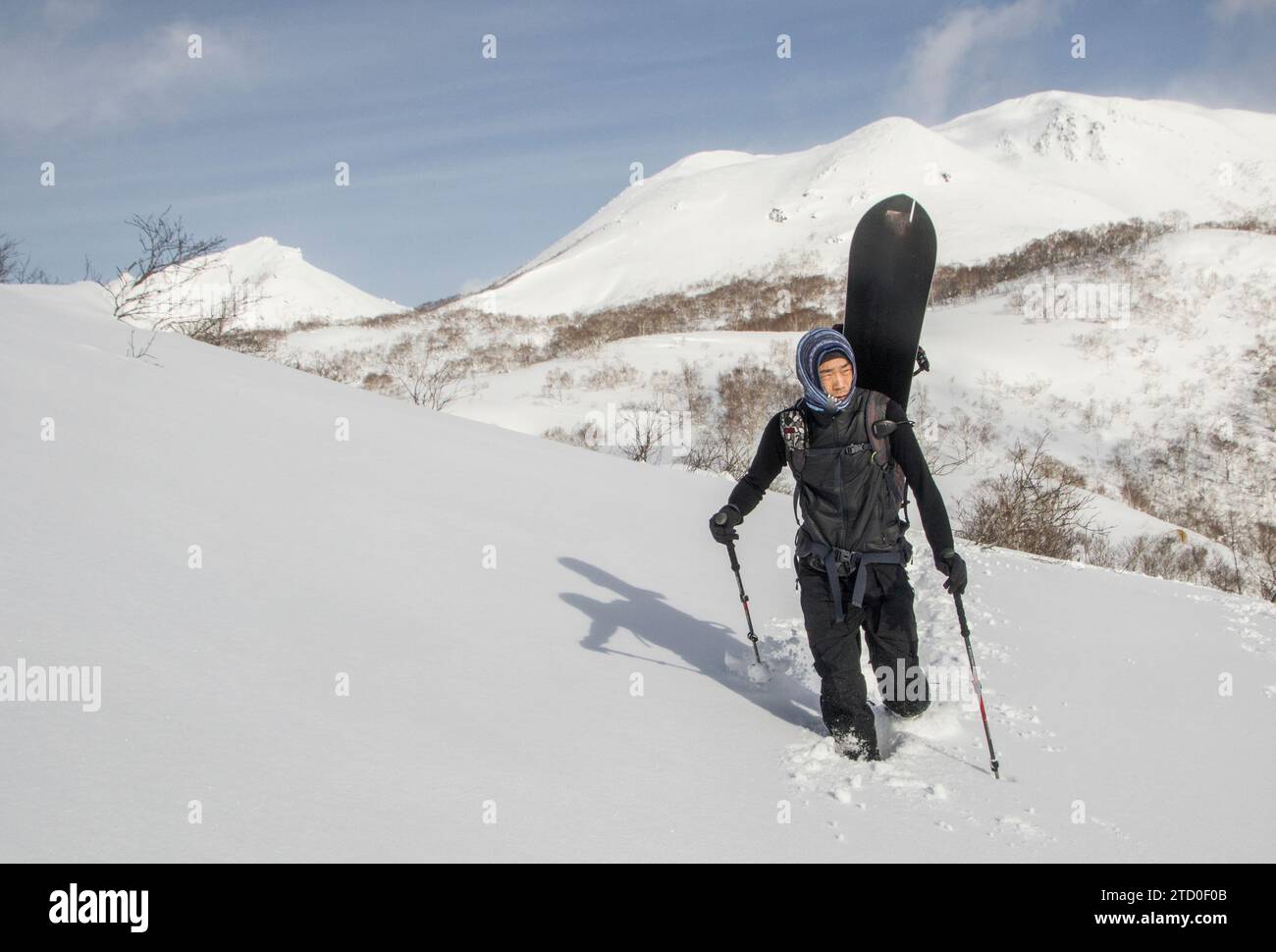 Tutto il corpo di un escursionista maschile in abiti caldi con snowboard e bastoncini che camminano sulle montagne innevate degli altopiani contro il cielo durante le vacanze invernali in Giappone Foto Stock