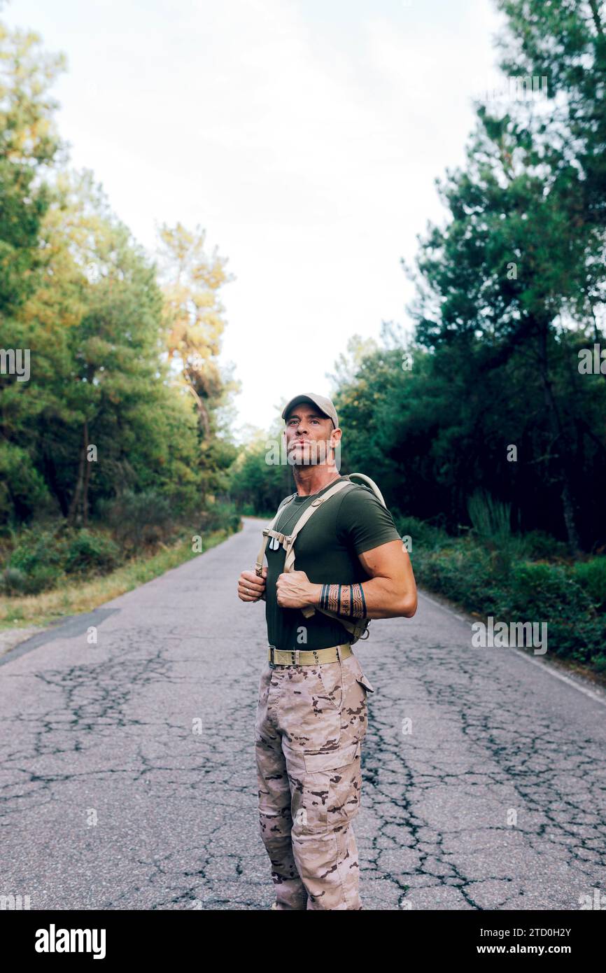 Soldato maturo e sicuro che indossa pantaloni camouflage e t-shirt verde che guarda lontano mentre si trova in strada contro gli alberi della foresta Foto Stock