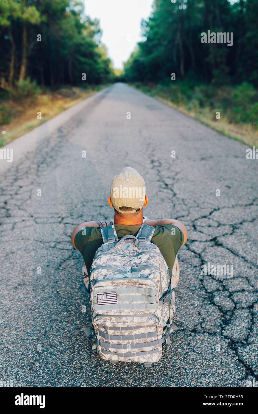 Alto angolo di anonimo soldato militare maschile che indossa un berretto con zaino mimetico seduto in strada contro gli alberi nella foresta Foto Stock