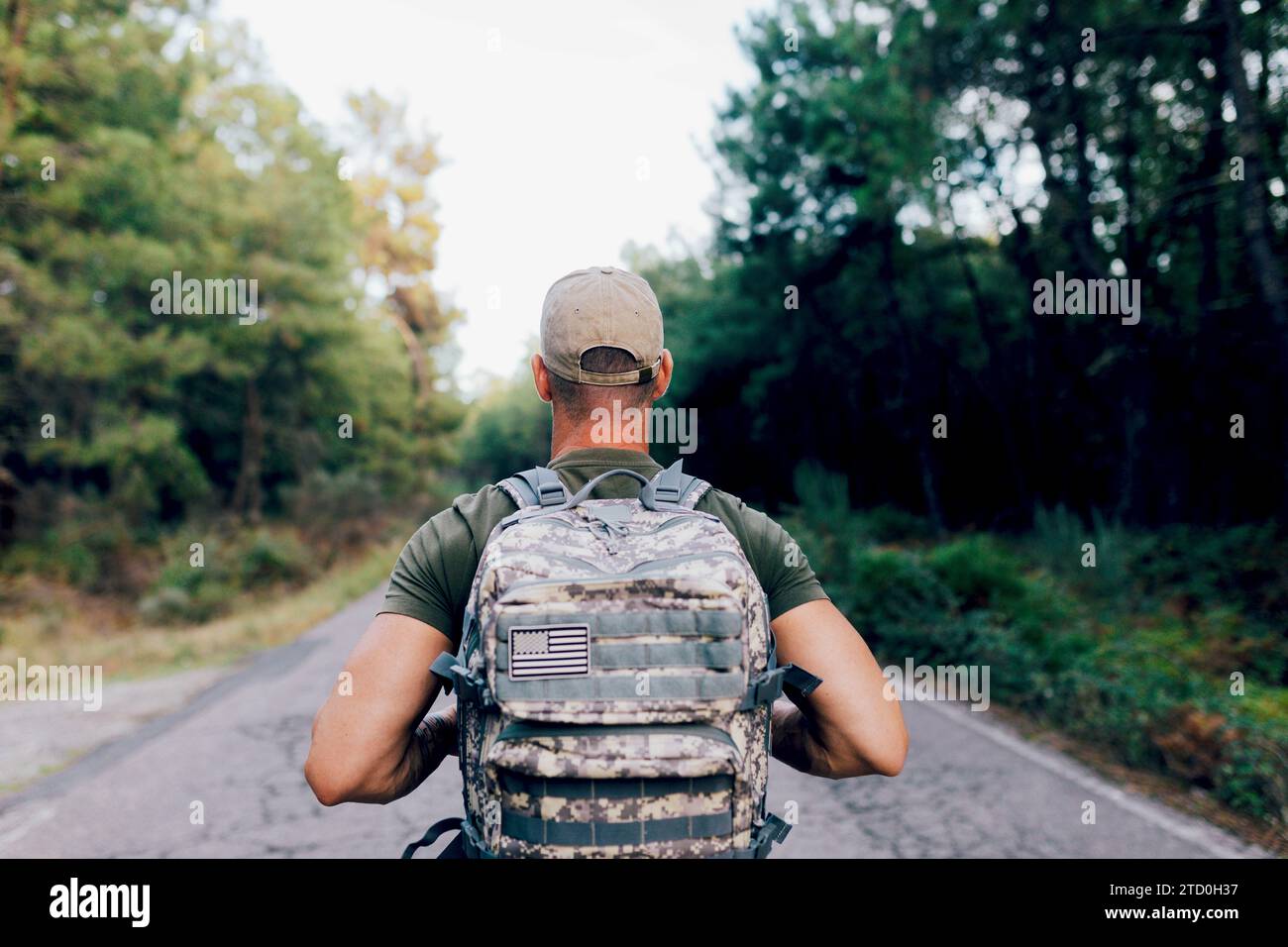 Vista posteriore di un soldato militare anonimo che indossa un berretto con uno zaino mimetico che ritorna mentre cammina sulla strada contro gli alberi nella foresta Foto Stock