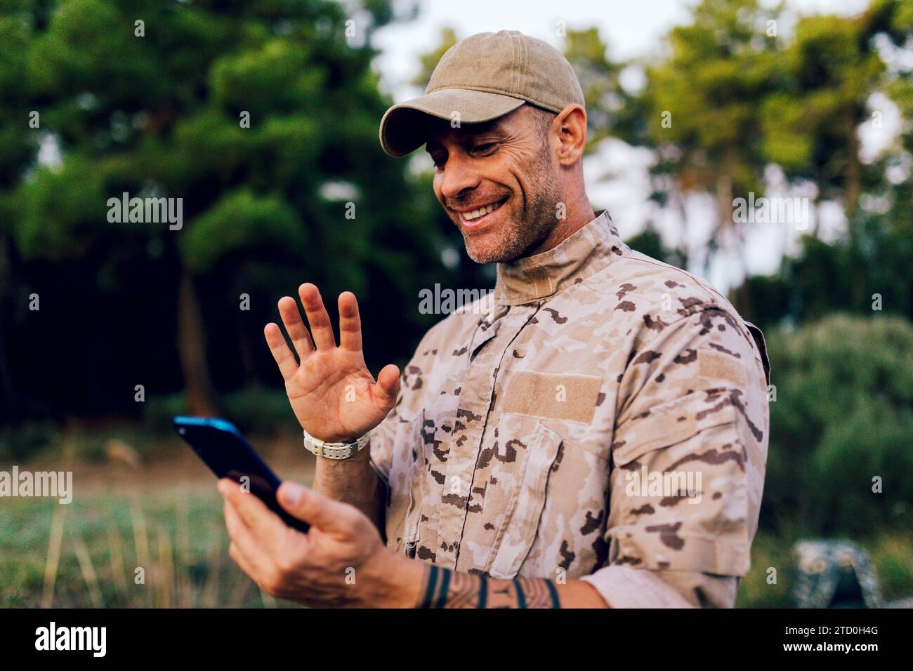 Felice soldato militare caucasico di mezza età che indossa un'uniforme mimetica che si muove allo smartphone durante le videochiamate nella foresta Foto Stock