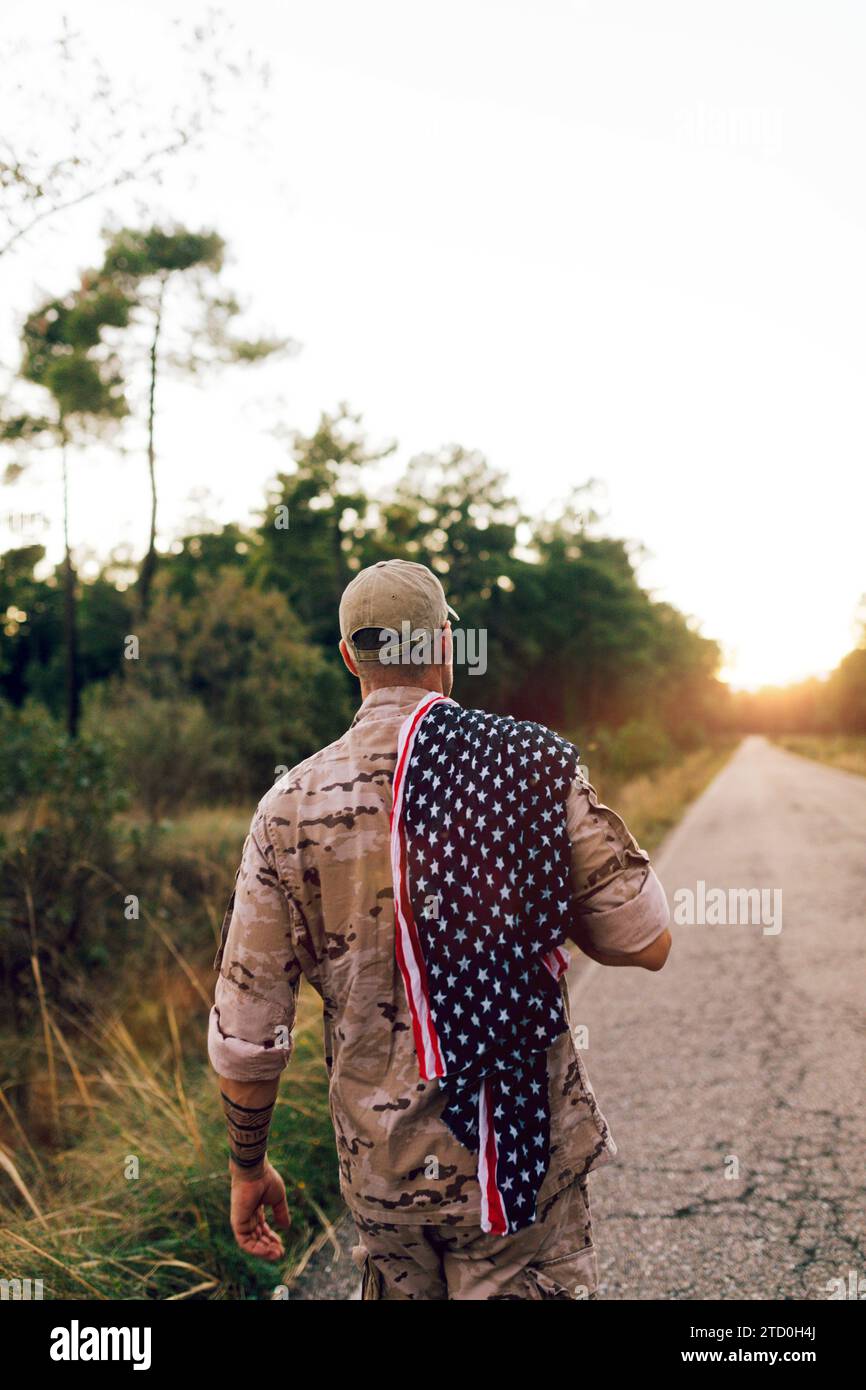 Vista posteriore di un soldato anonimo della forza militare speciale di mezza età che indossa un'uniforme mimetica che cammina su strada con la bandiera americana sulla spalla durante il sole Foto Stock