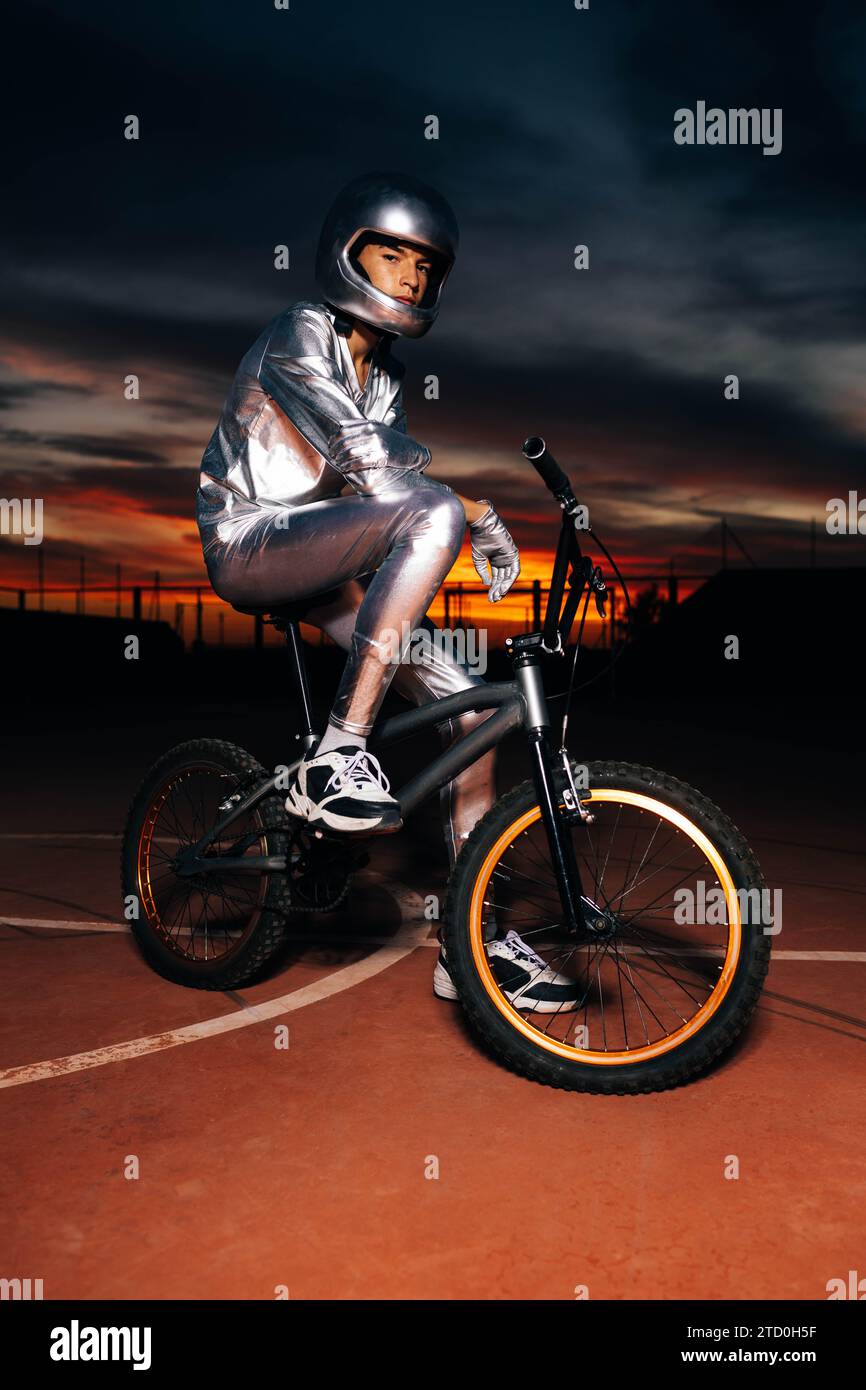 Vista laterale del giovane stuntman anonimo in costume argentato e casco che guarda la macchina fotografica seduto sul sedile della bicicletta con un piede da ginnastica sul campo sportivo Foto Stock