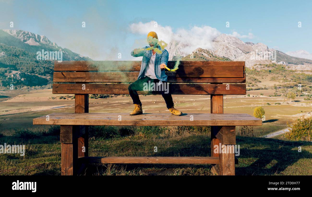 Una persona siede su una grande panchina di legno circondata da montagne, rilasciando una nuvola di fumo colorato e vibrante. Foto Stock