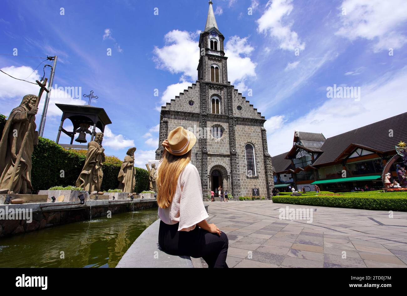 Giovane donna turistica elegante con cappello seduto a parete che guarda la chiesa madre di Gramado, famoso punto di riferimento e destinazione di viaggio nello stato del Rio grande do sul, Foto Stock
