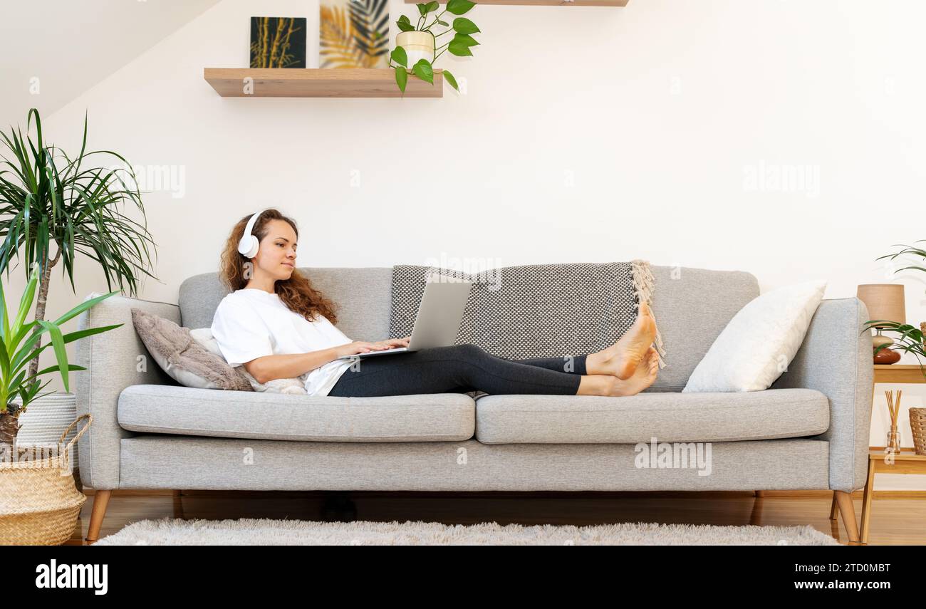 Donna con cuffie wireless sdraiata sul divano e che usa il computer portatile. Foto Stock