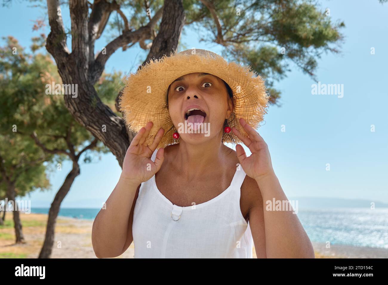 Attraente donna sorridente di mezza età che indossa un cappello di paglia al mare. Donna matura felice che si gode la libertà in mare e fa volti che guardano la fotocamera. Foto Stock