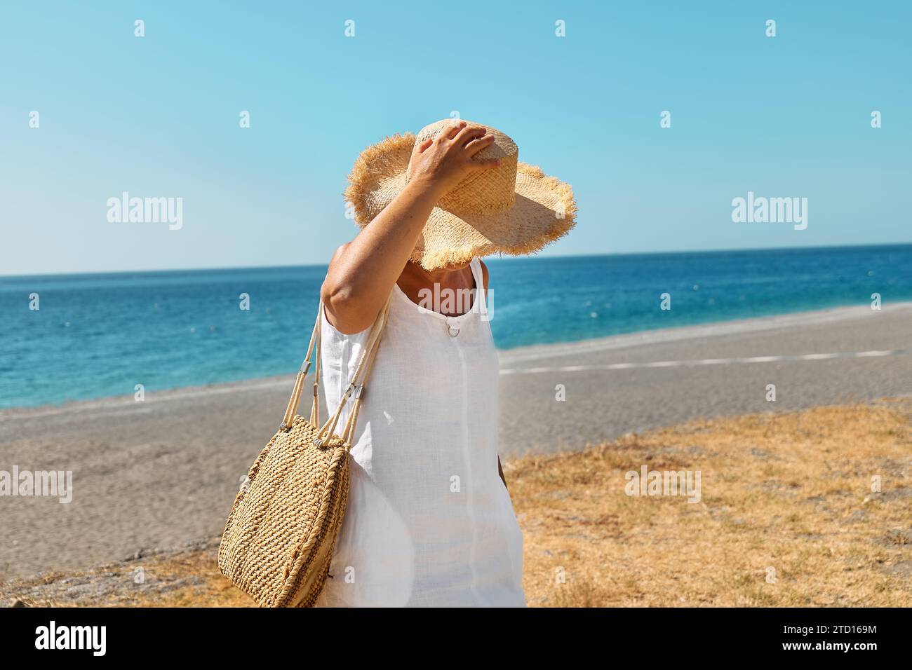 Vista laterale dell'attraente donna di mezza età che indossa un cappello di paglia al mare. Donna matura felice che cammina in spiaggia, godendo della libertà in mare. Foto Stock