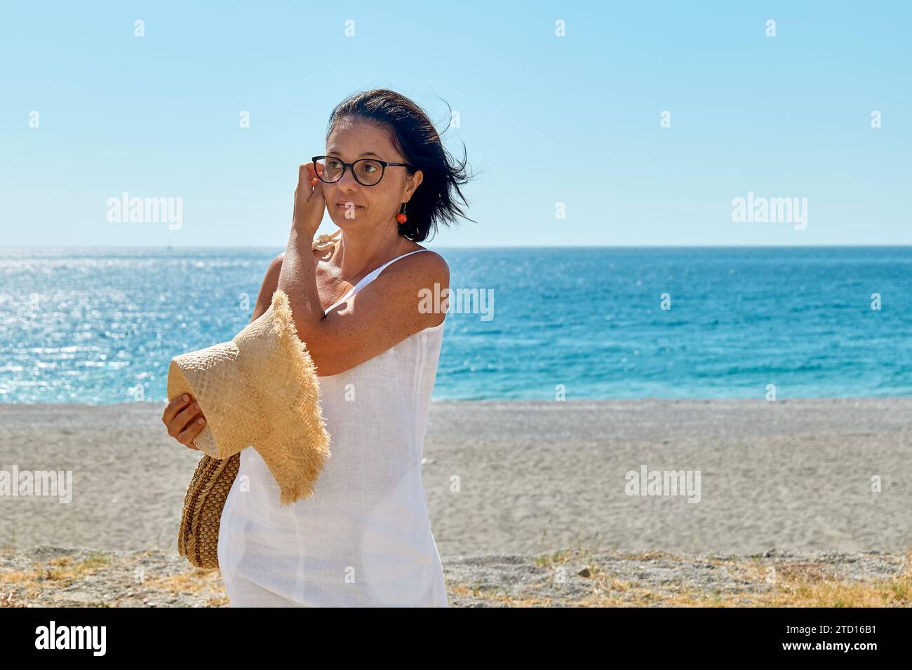 Attraente donna di mezza età che indossa un cappello di paglia al mare. Donna matura felice che cammina in spiaggia, godendo della libertà in mare. Foto Stock