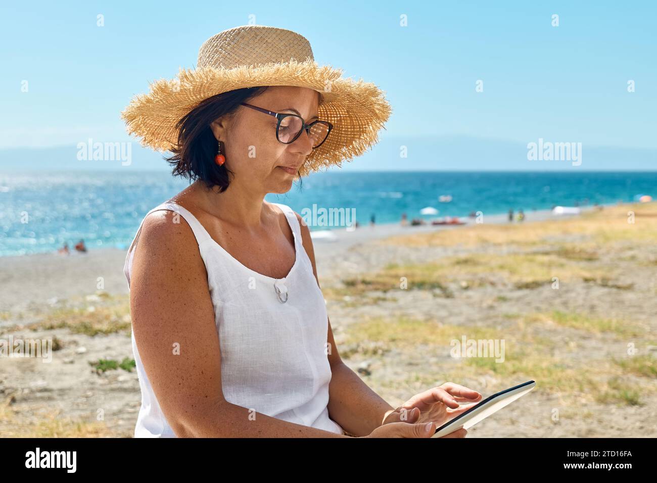 Donna matura che indossa un cappello di paglia utilizzando un tablet digitale in spiaggia. Donna di mezza età che utilizza un dispositivo digitale per lo shopping online, bancario, social media o Foto Stock