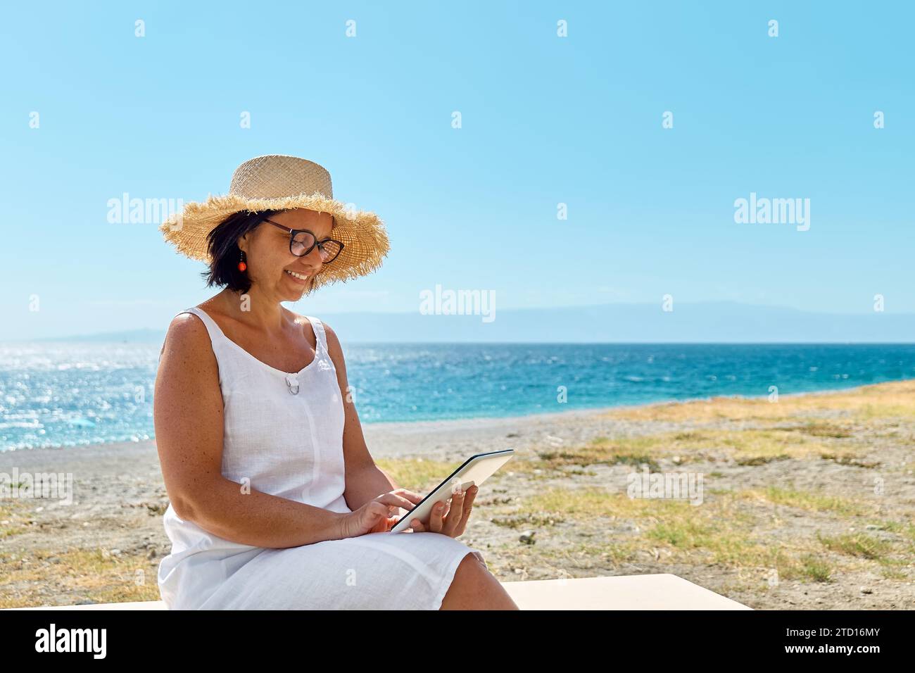 Donna matura che indossa un cappello di paglia utilizzando un tablet digitale in spiaggia. Donna di mezza età che utilizza un dispositivo digitale per lo shopping online, bancario, social media o Foto Stock