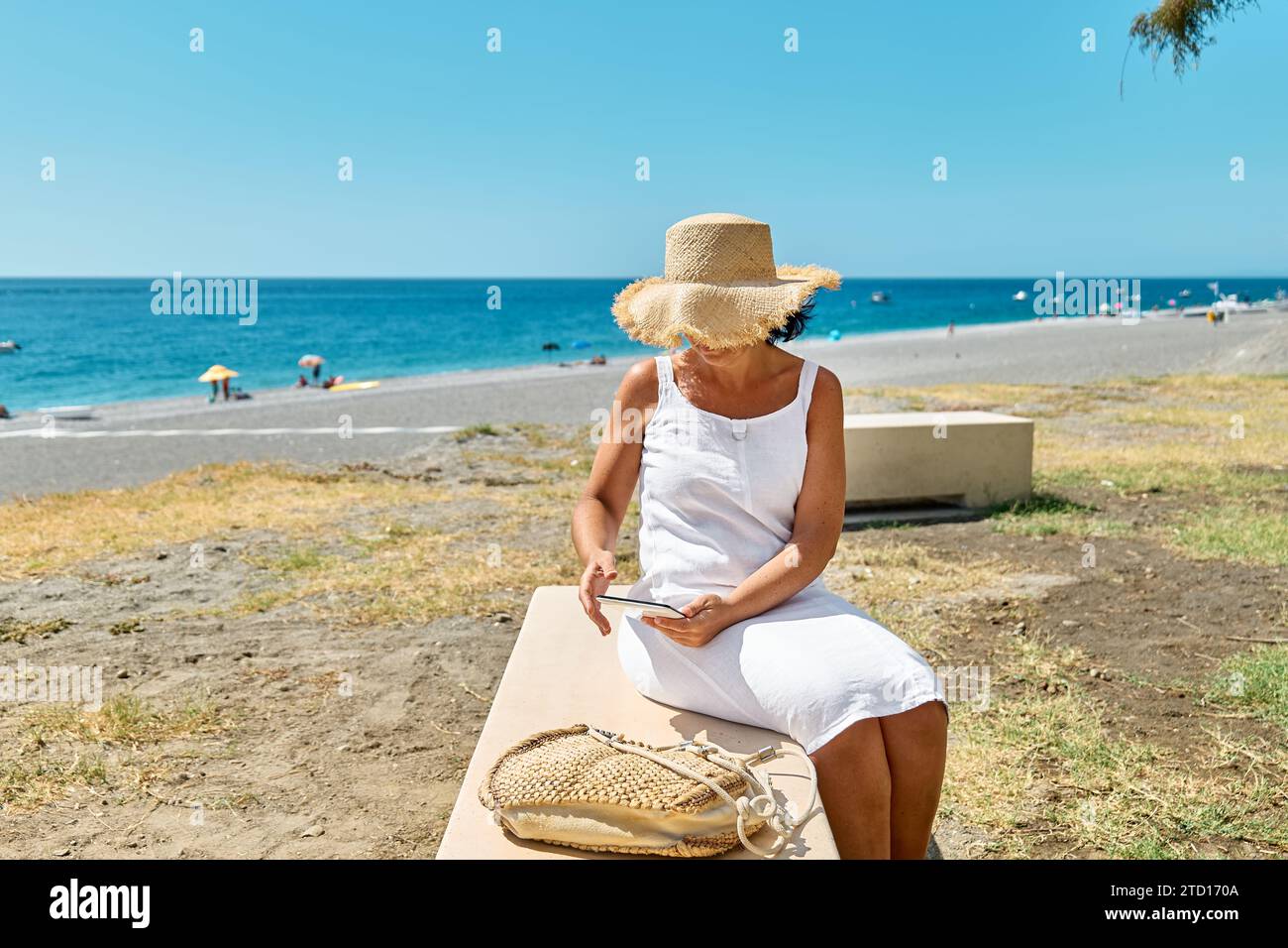 Donna matura che indossa un cappello di paglia utilizzando un tablet digitale in spiaggia. Donna di mezza età che utilizza un dispositivo digitale per lo shopping online, bancario, social media o Foto Stock