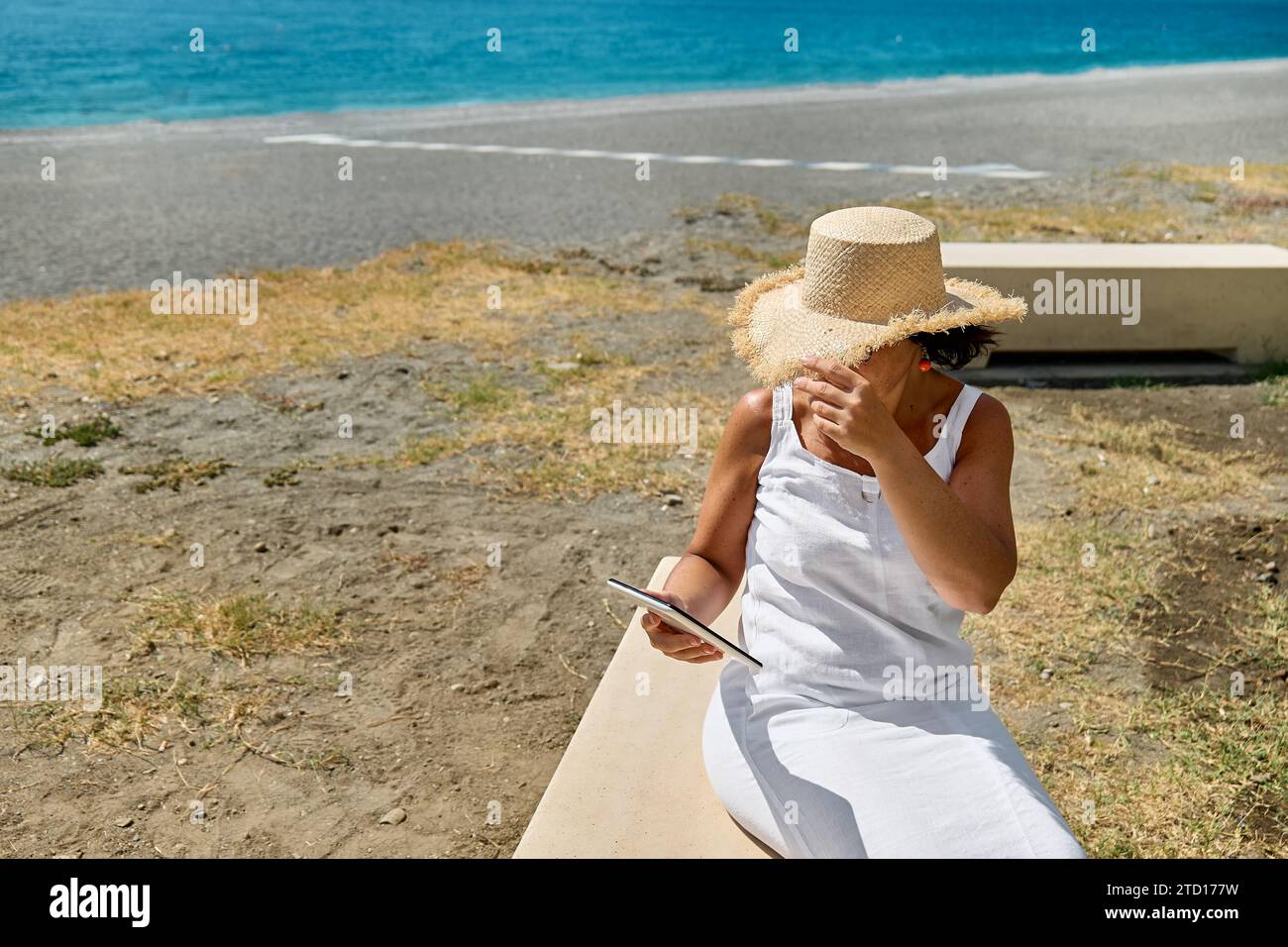 Donna matura che indossa un cappello di paglia utilizzando un tablet digitale in spiaggia. Donna di mezza età che utilizza un dispositivo digitale per lo shopping online, bancario, social media o Foto Stock