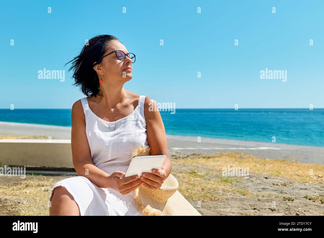 Donna matura che indossa un tablet digitale in spiaggia. Donna di mezza età che utilizza un dispositivo digitale per lo shopping online, bancario, sui social media o per la lettura Foto Stock
