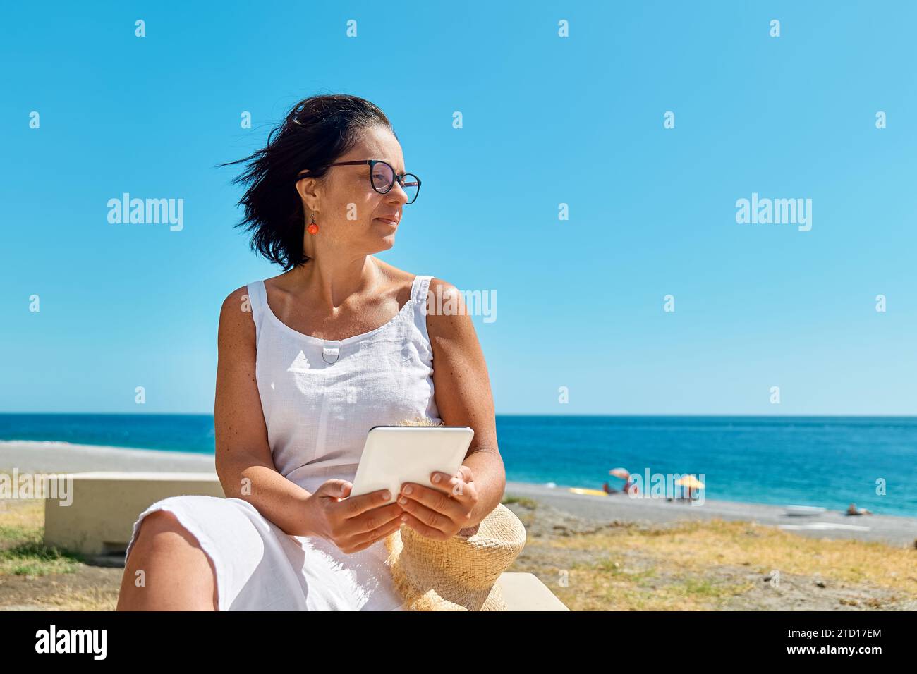 Donna matura che indossa un tablet digitale in spiaggia. Donna di mezza età che utilizza un dispositivo digitale per lo shopping online, bancario, sui social media o per la lettura Foto Stock
