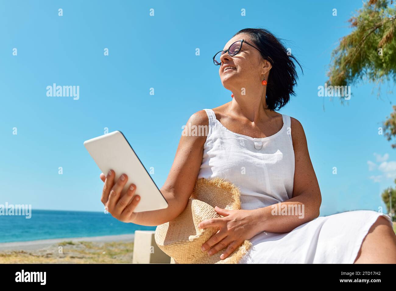 Donna matura che indossa un tablet digitale in spiaggia. Donna di mezza età che utilizza un dispositivo digitale per lo shopping online, bancario, sui social media o per la lettura Foto Stock