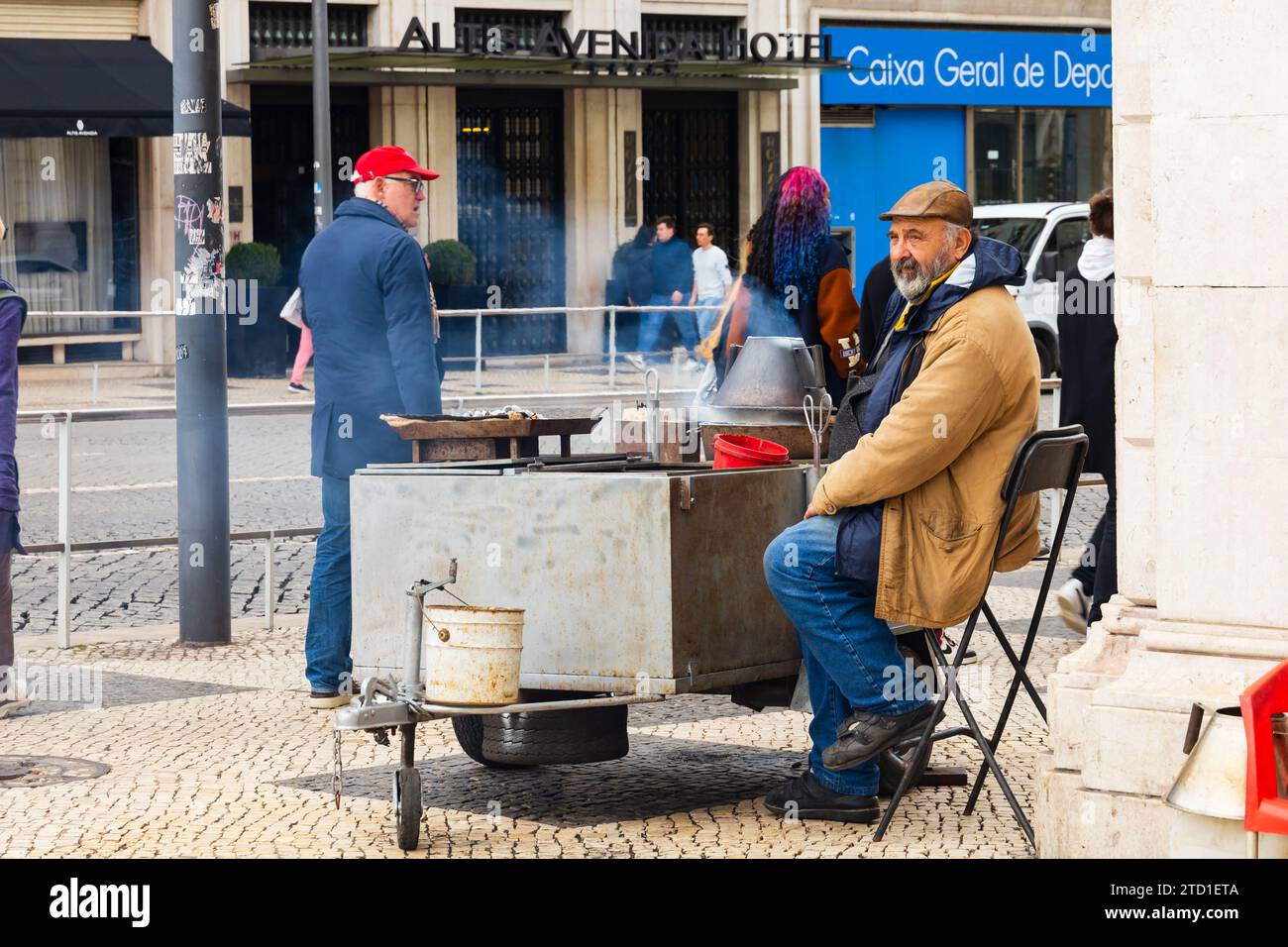 I pedoni attraversano una strada Vendor che vende le tradizionali castagne arrosto dal suo fornello per le strade di Lisbona, Portogallo Foto Stock