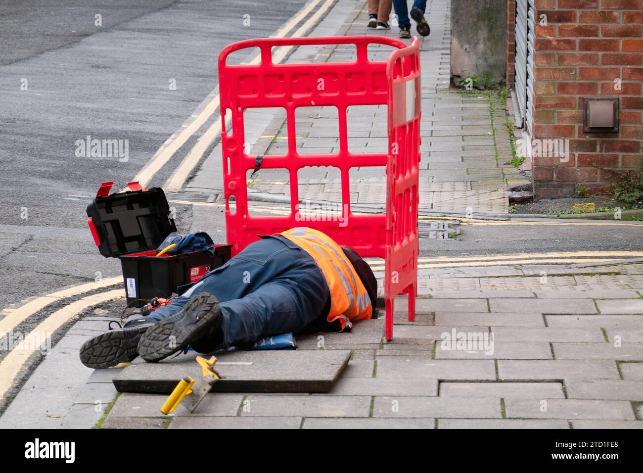 Un ingegnere BT Openreach sdraiato su un marciapiede che lavora su cavi telefonici sotterranei a cui si accede sollevando una copertura di ispezione su una strada Foto Stock
