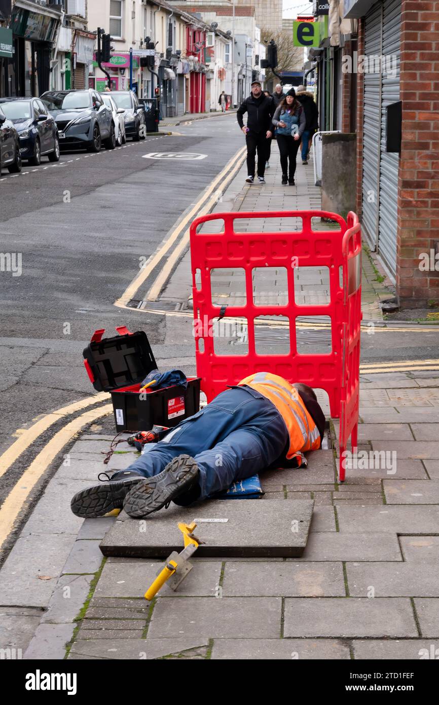 Un ingegnere BT Openreach sdraiato su un marciapiede che lavora su cavi telefonici sotterranei a cui si accede sollevando una copertura di ispezione su una strada Foto Stock