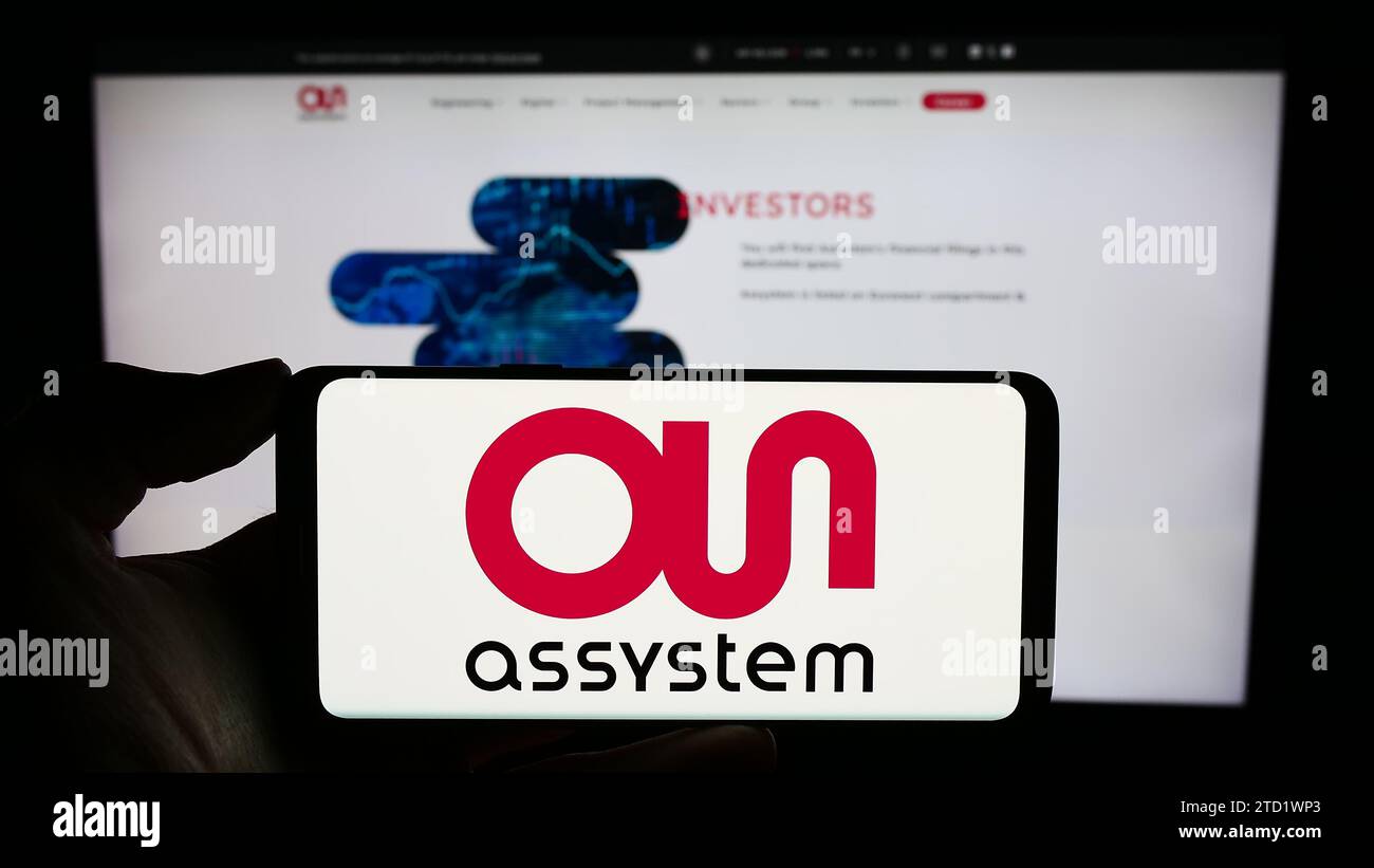 Persona che possiede il cellulare con il logo della società di ingegneria francese Assystem S.A. davanti alla pagina Web aziendale. Concentrarsi sul display del telefono. Foto Stock