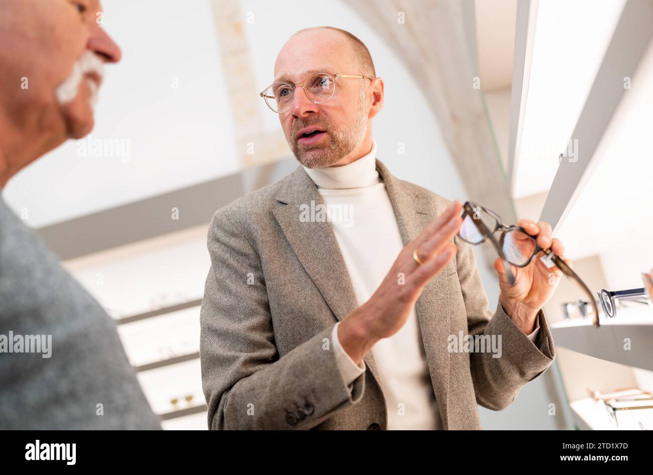 Un ottico che parla di occhiali da vista con un cliente senior in negozio. Sta indicando gli occhiali. Foto Stock