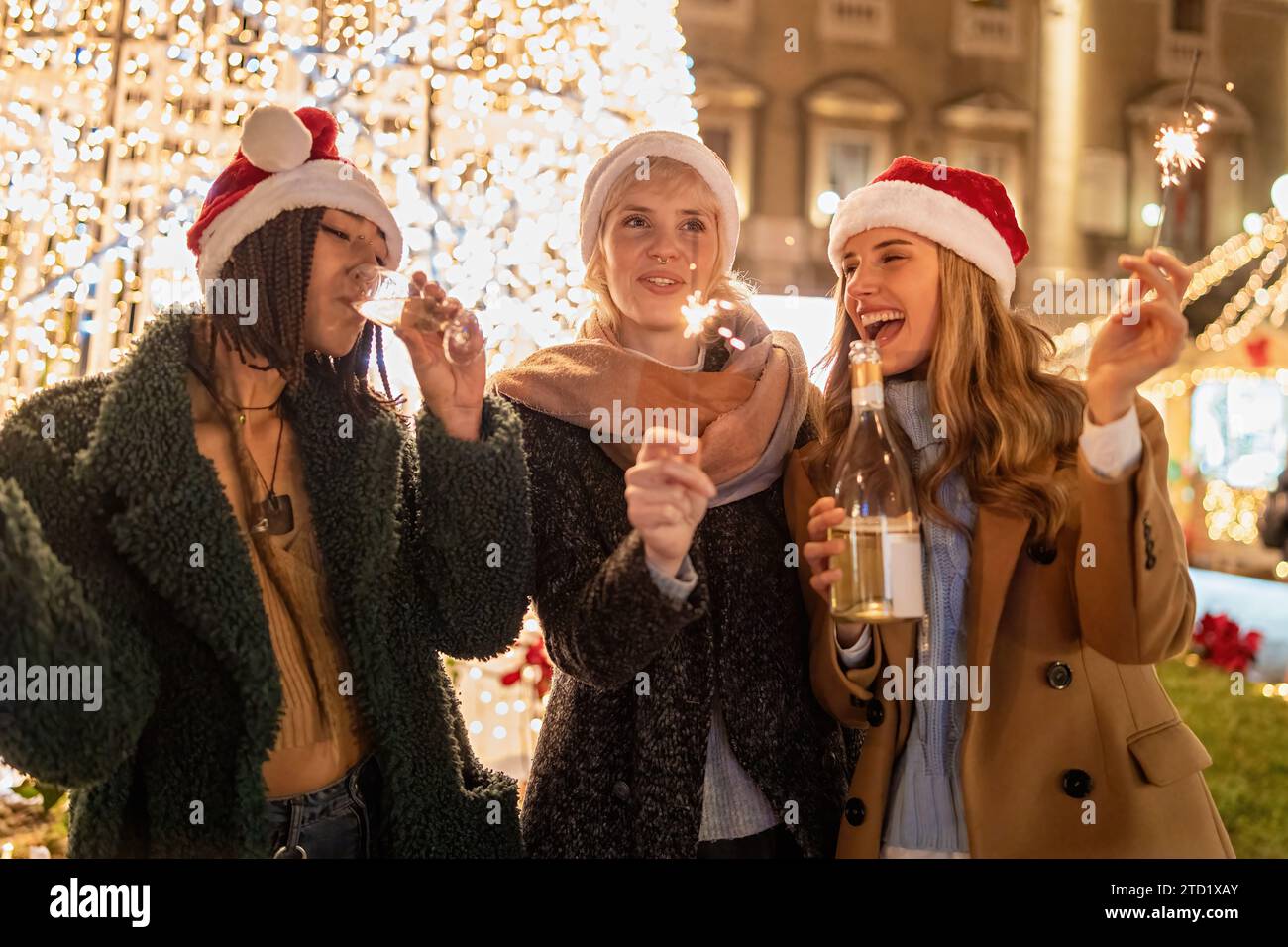 Tre donne condividono risate e scintillanti, abbracciando lo spirito festivo con cappelli di Natale e una bottiglia di vino, illuminati dalle luci della città. Foto Stock