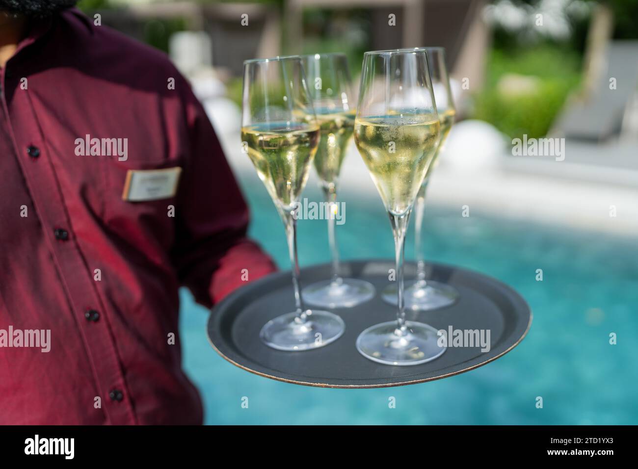 Cameriere con una camicia rossa che tiene in mano un vassoio con tre bicchieri di champagne a bordo piscina. Immagine del concetto di viaggio alberghiero Foto Stock