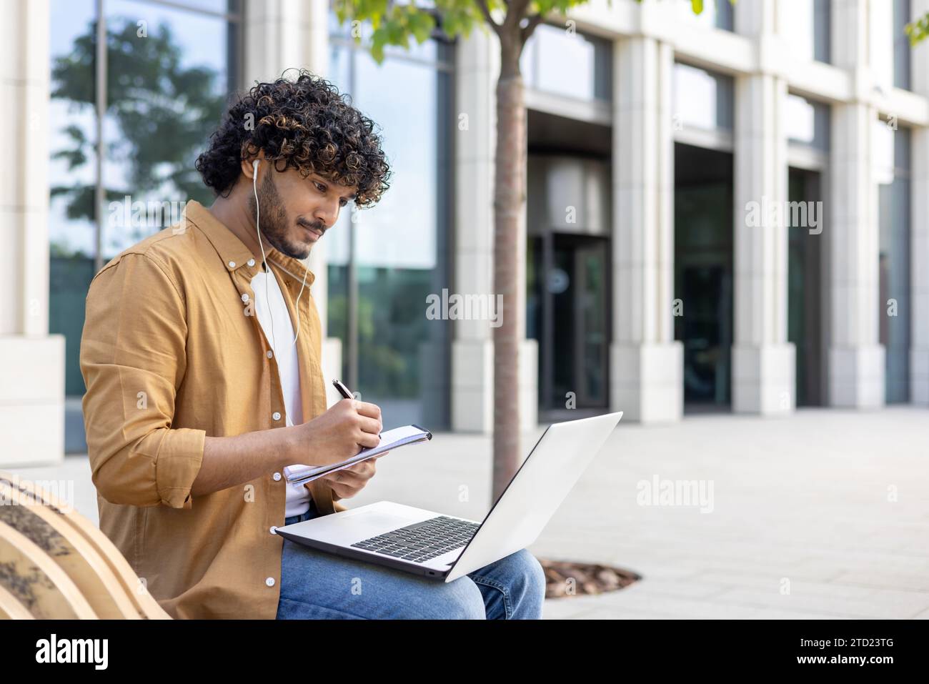 Giovane studente maschio indiano seduto fuori su una panchina e studiando online, guardando e ascoltando in cuffia le lezioni online, scrivendo in un n Foto Stock