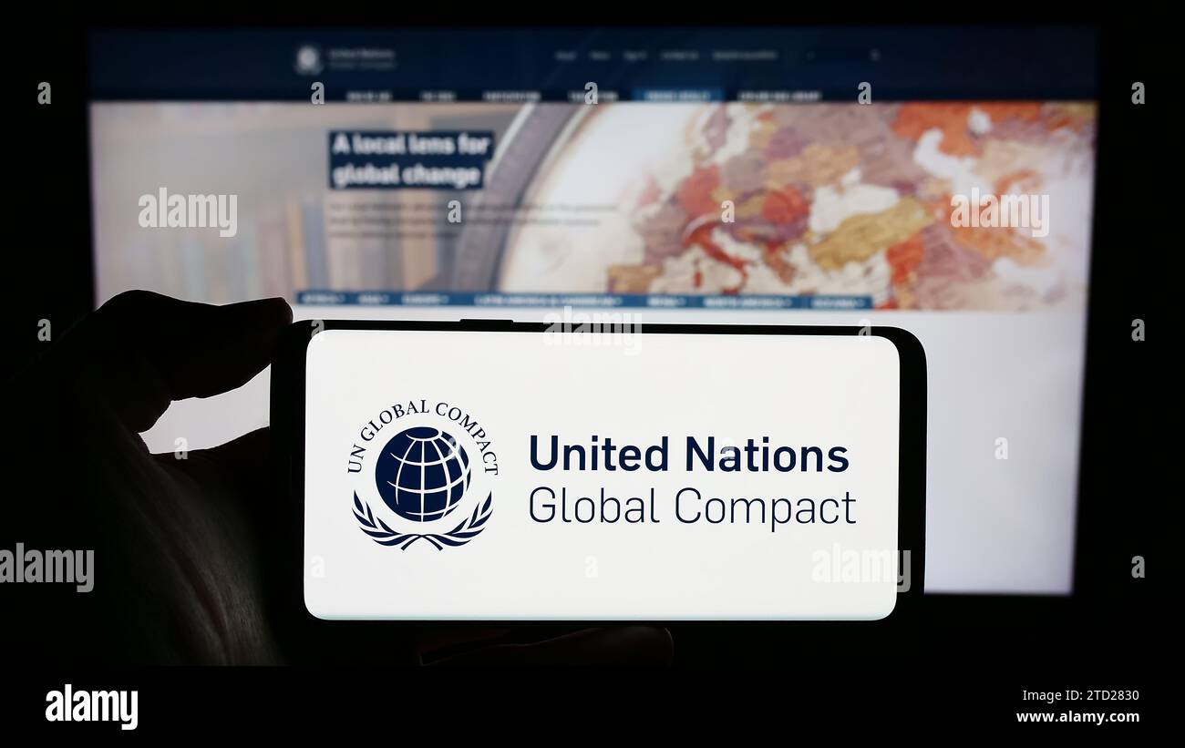 Persona in possesso di un cellulare con il logo del Patto delle Nazioni Unite Global Compact davanti alla pagina web. Concentrarsi sul display del telefono. Foto Stock