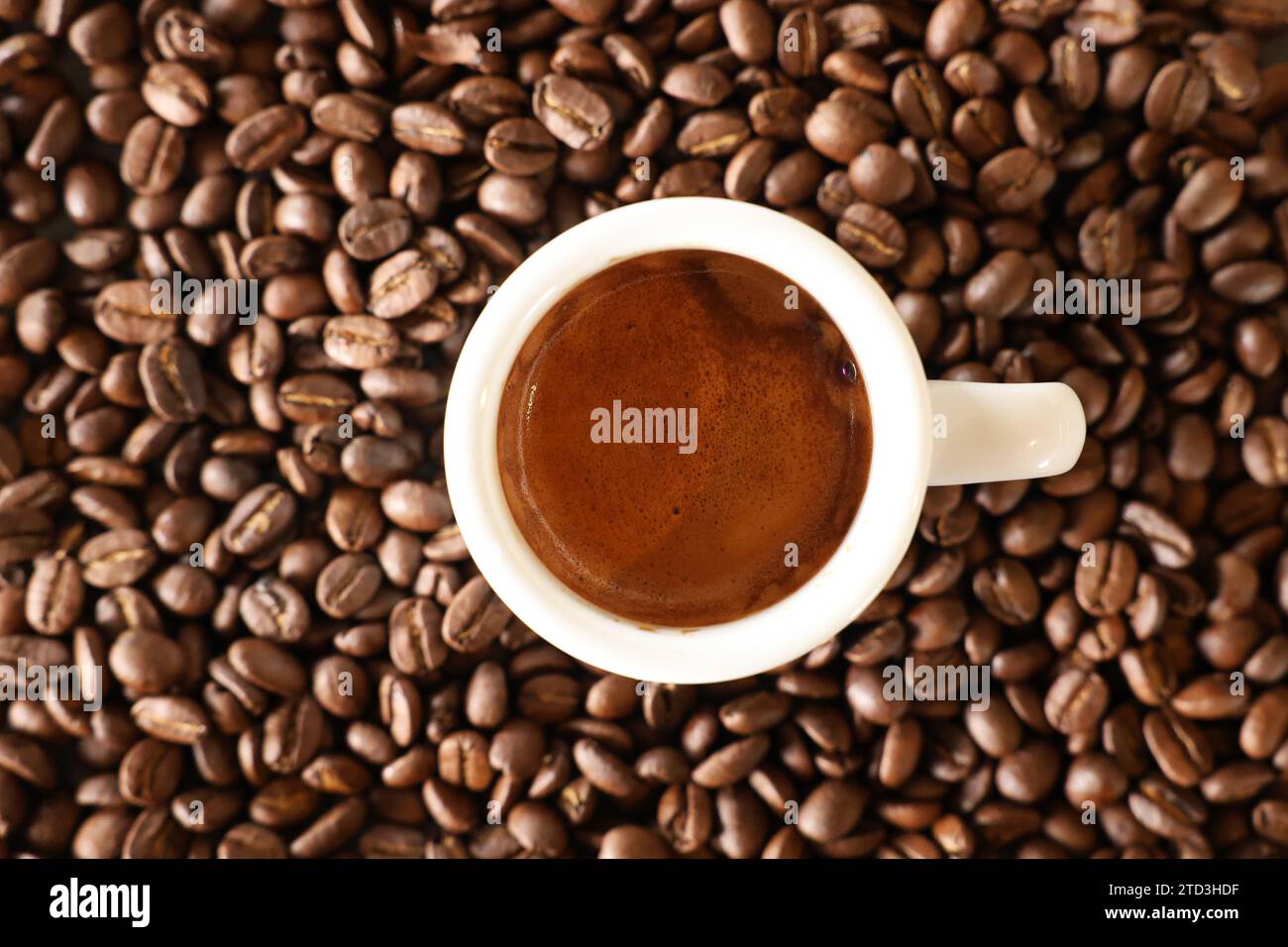 Una vista che guarda direttamente in basso su una tazza di caffè bianco riempita con una tazza di caffè appena fatto con crema marrone. Sfondo di chicchi di caffè tostati. Foto Stock