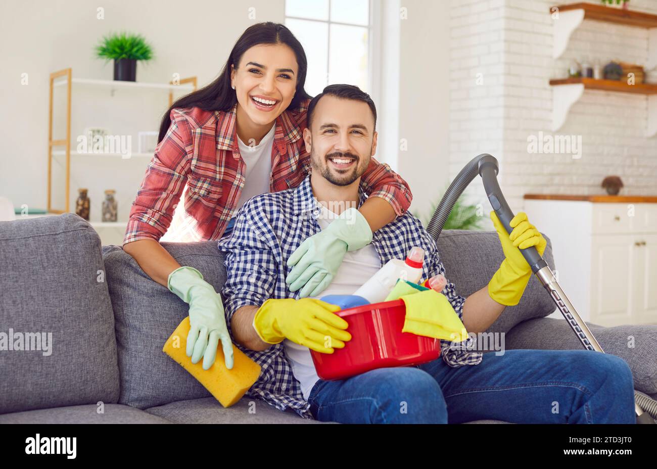 Coppia felice con i prodotti per la pulizia della casa in mano seduti sul divano a casa Foto Stock