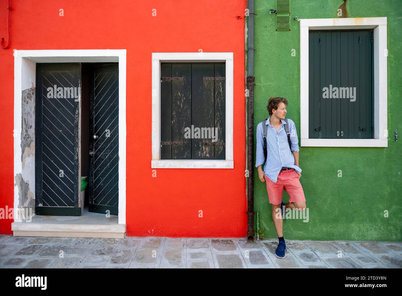 Giovane uomo con zaino appoggiato contro un muro di casa, facciata verde e rossa con porta d'ingresso e finestra, case colorate sull'isola di Foto Stock