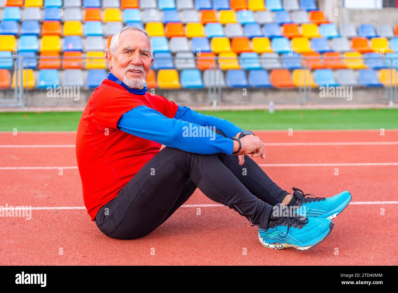 Ritratto di un uomo anziano con abiti sportivi seduto su una pista atletica e sorridente davanti alla fotocamera che sembra stanco Foto Stock