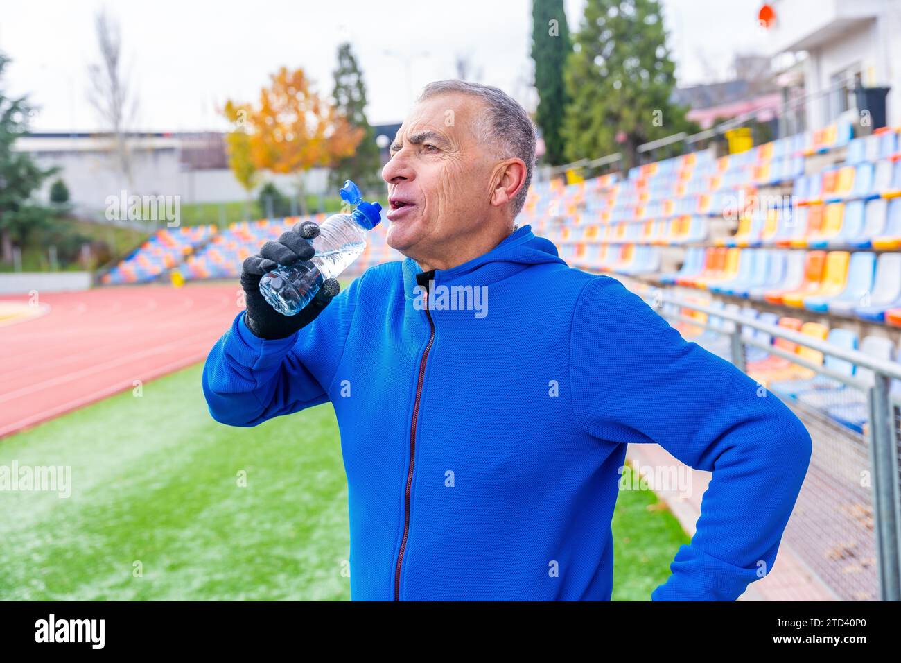 Uomo maturo con abiti sportivi blu che beve acqua dopo aver corso in pista all'aperto in inverno Foto Stock