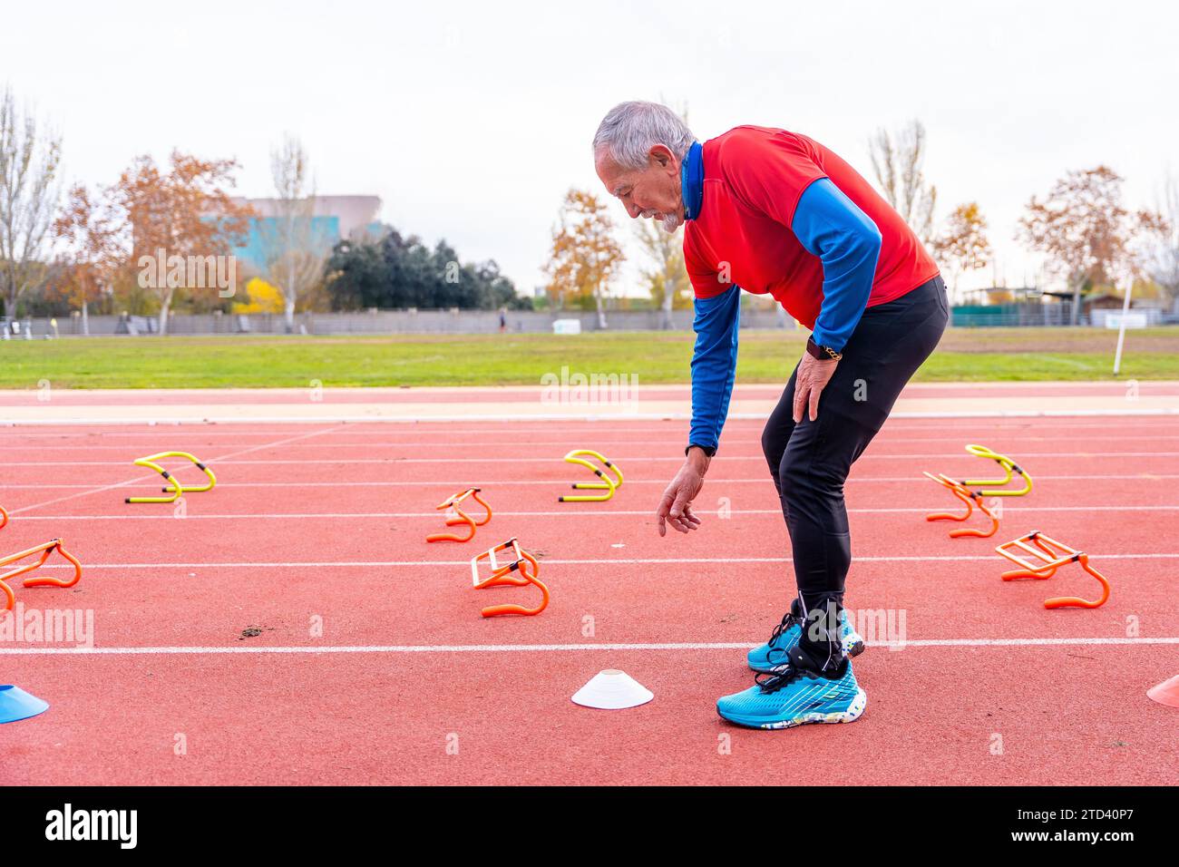 Uomo anziano sportivo che posiziona coni e blocchi di partenza su una pista di atletica per allenarsi all'aperto in inverno Foto Stock