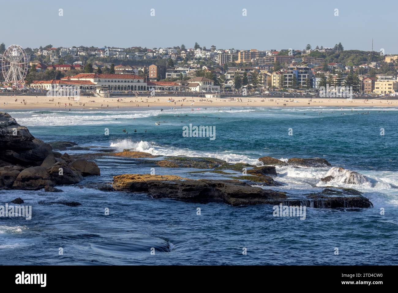 Onde che si innalzano sulle rocce in primo piano, prima dell'iconica Bondi Beach e del Bondi Pavilion, a Sydney, Australia. Le persone si godono la sabbia e l'acqua. Foto Stock