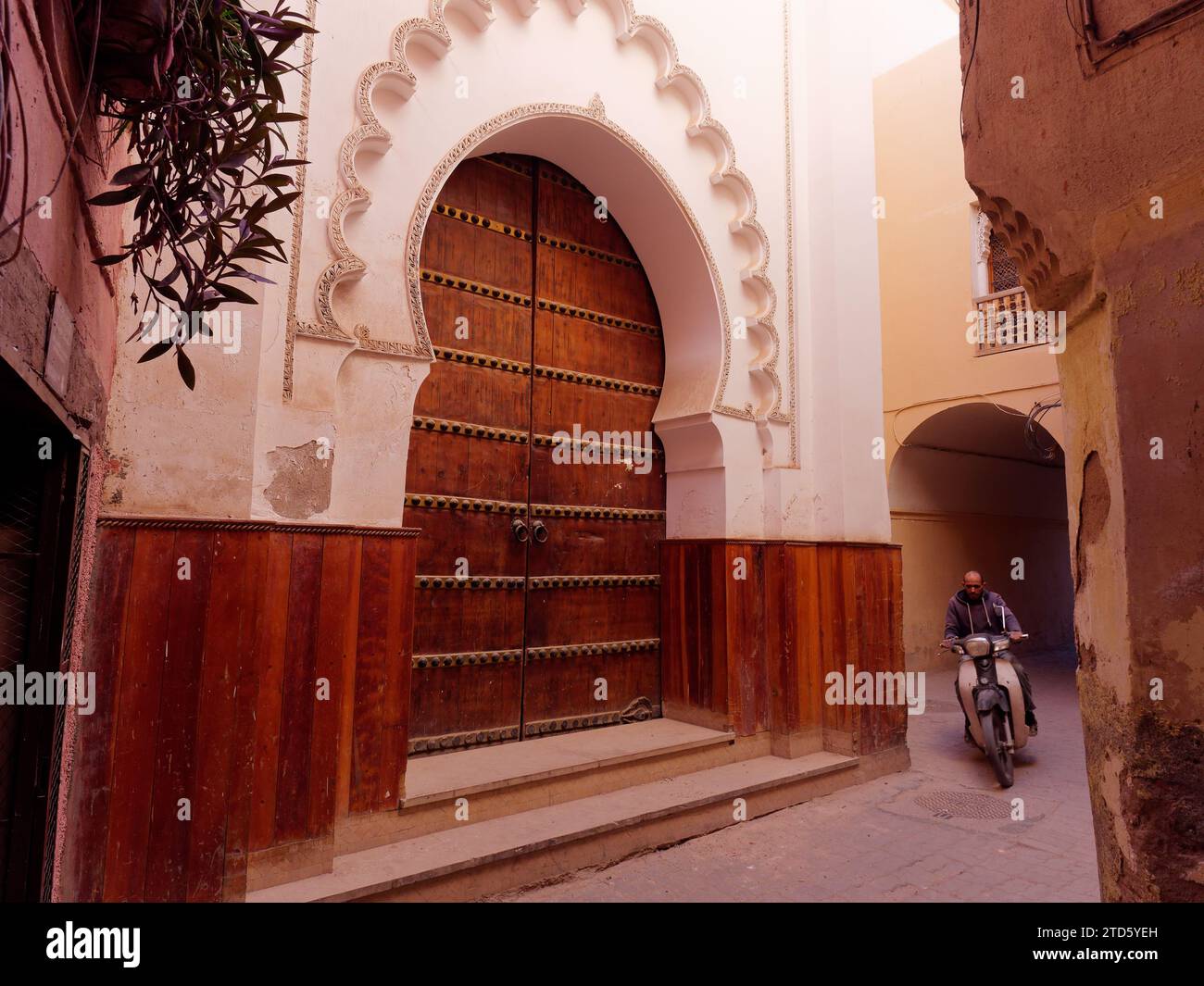 Scooter rider ed elegante grande porta d'ingresso in legno in una strada nella Medina di Marrakech durante il giorno, Marocco, 16 dicembre 2023 Foto Stock